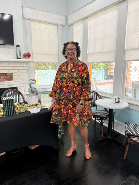 Woman in colorful dress standing in a room with large windows, a table, and chairs.