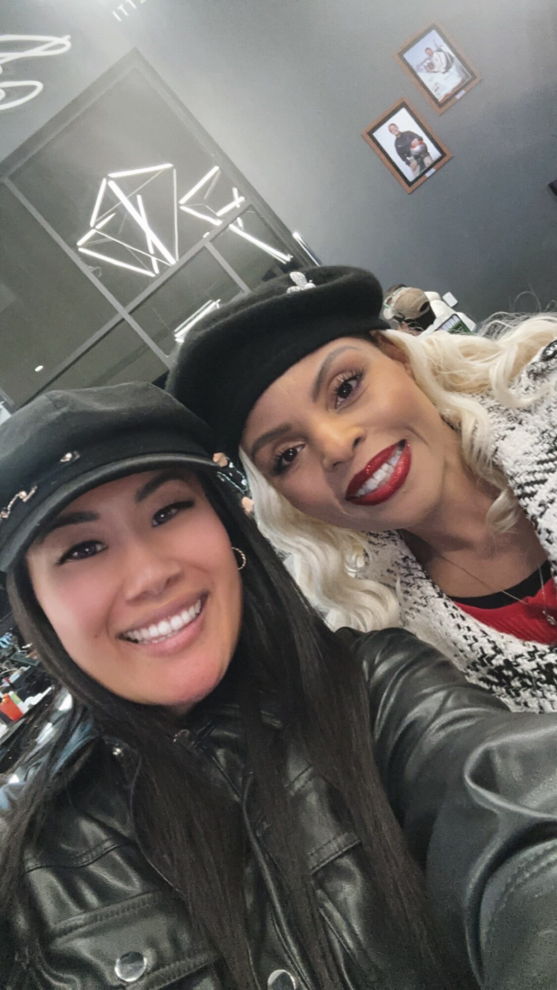 Two women smiling, wearing black hats and leather jackets, in an indoor setting with framed pictures on the wall.