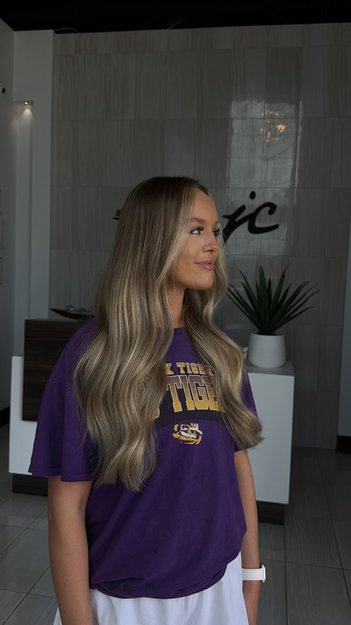 Young woman with long, wavy hair wearing a purple T-shirt, standing indoors near a plant and a wall with text.