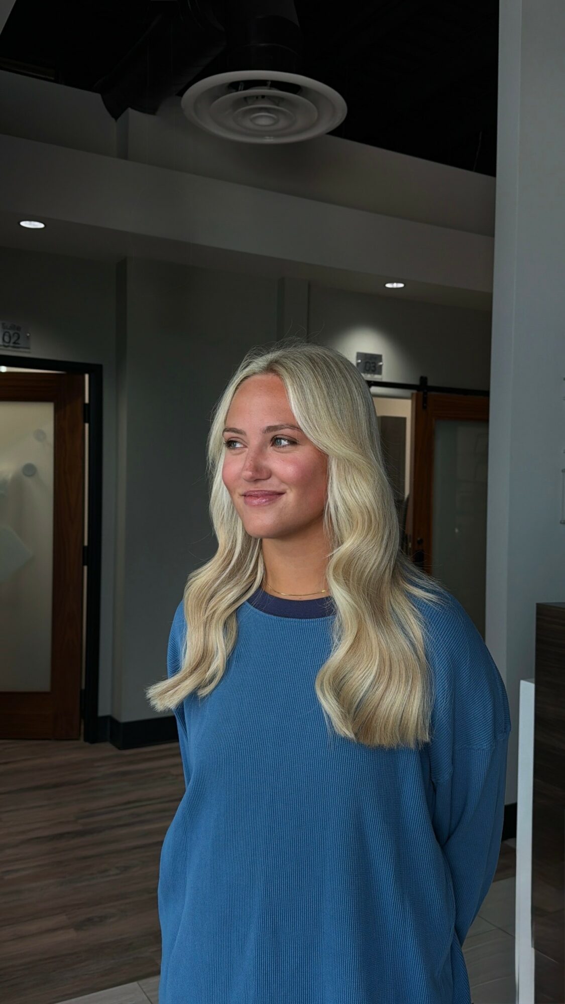 Young woman with long, wavy blonde hair wearing a blue sweater, standing indoors with a neutral expression.