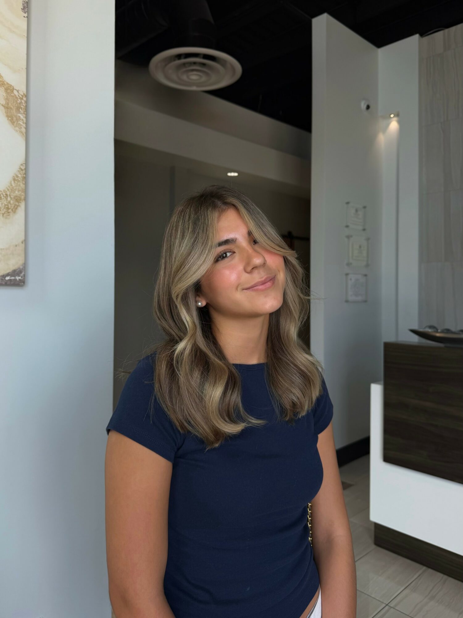 Young woman with wavy blonde hair smiling, wearing a navy blue shirt, indoors with modern decor.