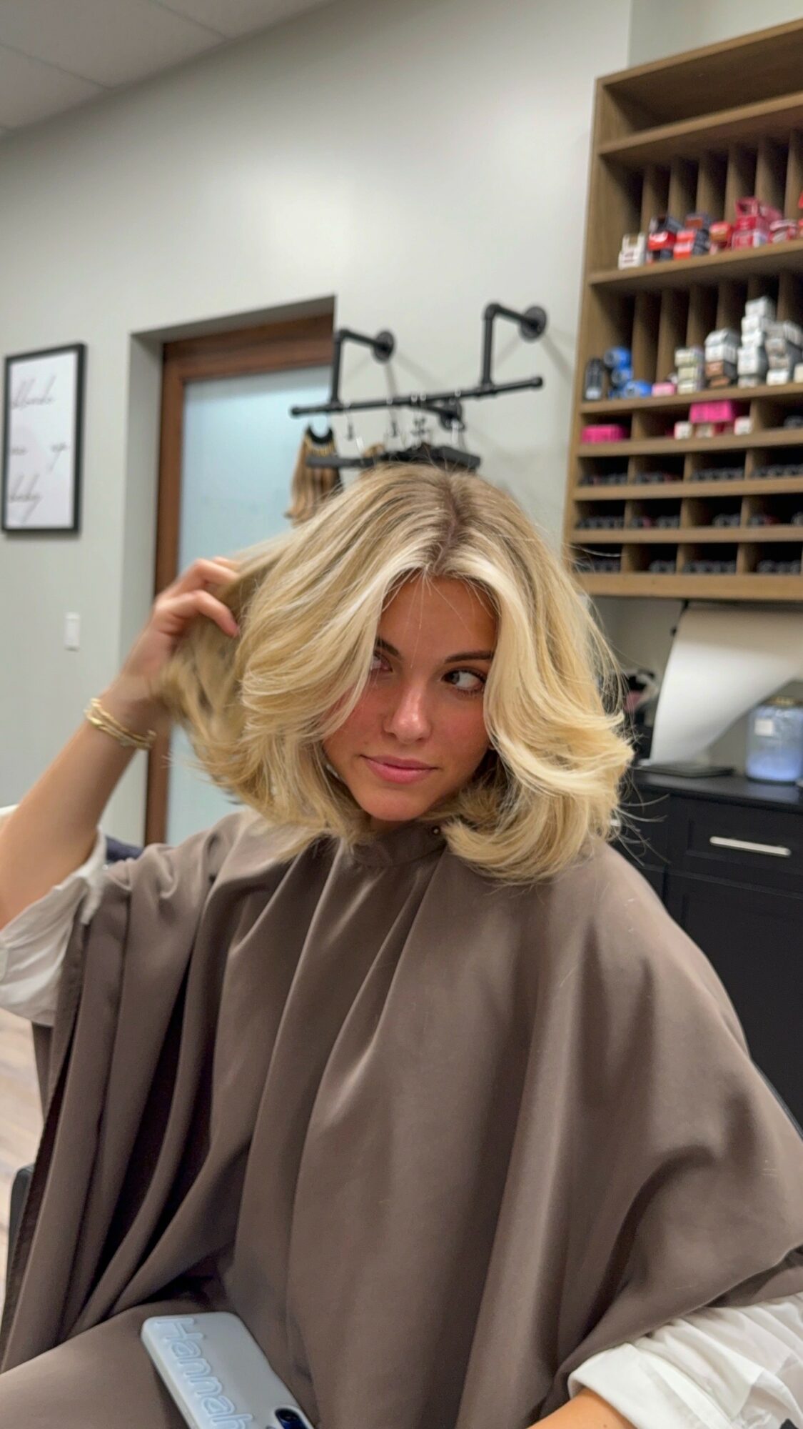 Woman with blonde, wavy hair in a salon chair, wearing a brown cape, in a salon with shelves of nail polish.