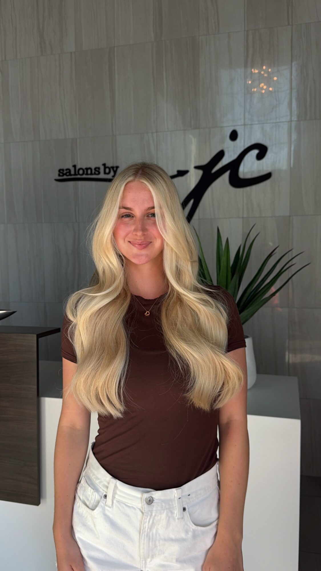 Young woman with long, wavy blonde hair smiling, wearing a brown top and white pants, standing indoors.
