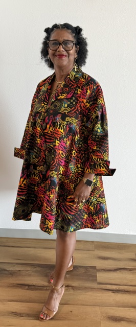 Woman with curly hair and glasses wearing a colorful patterned dress, standing indoors against a white wall.