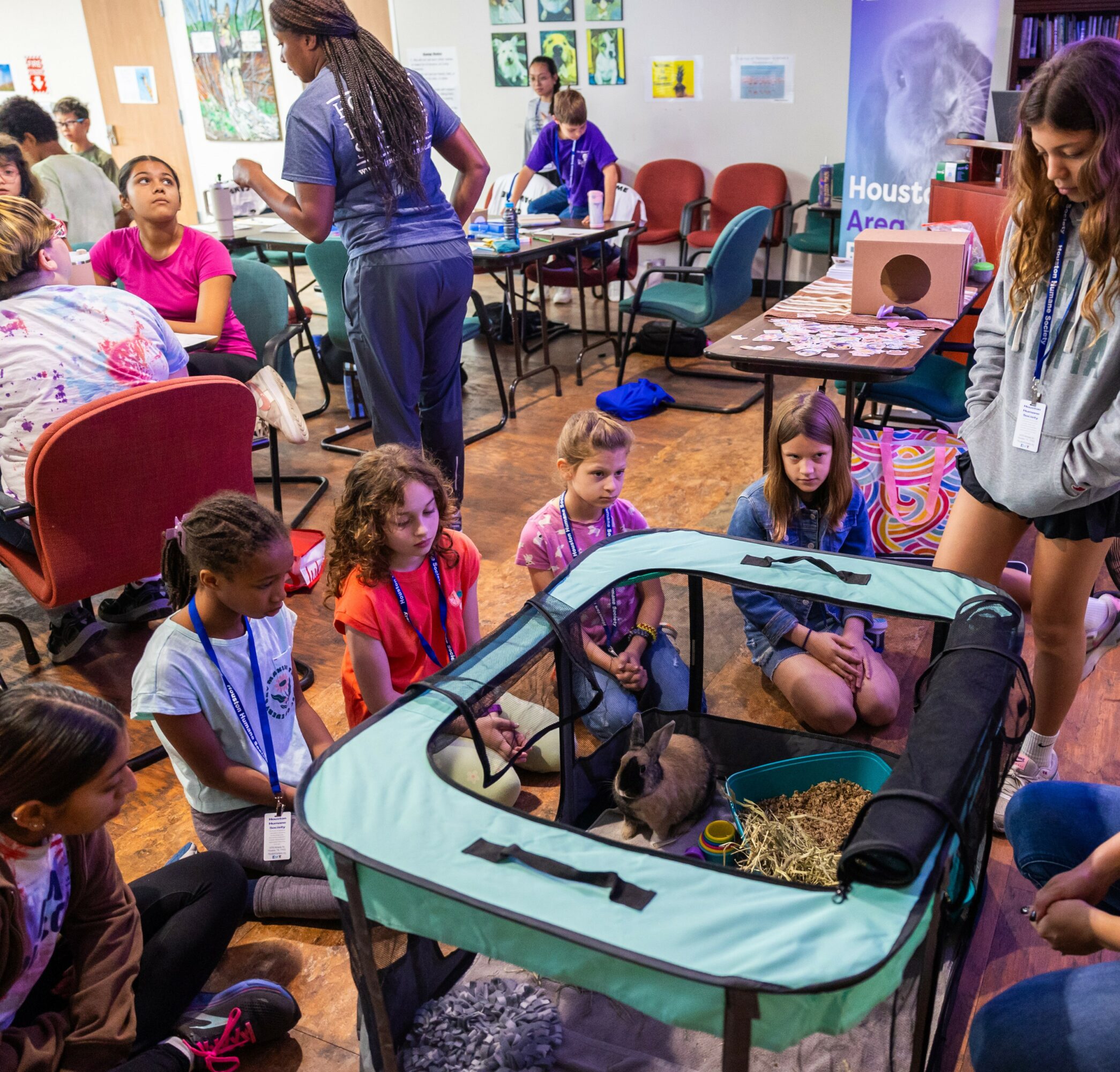 Children and adults gather around a baby in a portable crib with a mesh side, in a room with chairs and tables.