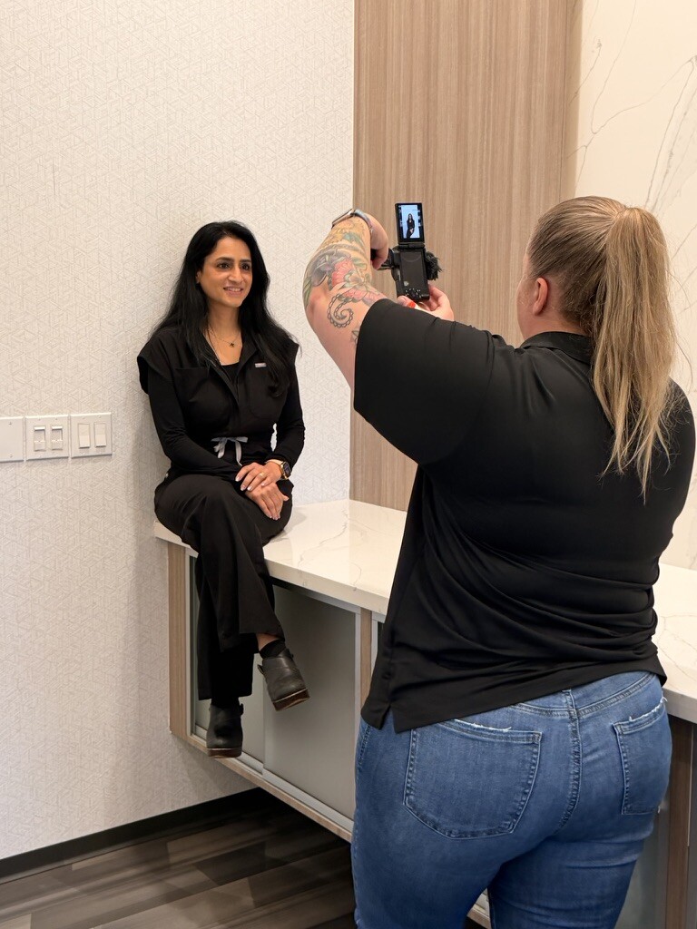 Woman sitting on a small ledge, posing for a photo taken by another woman with a smartphone, in an indoor setting.