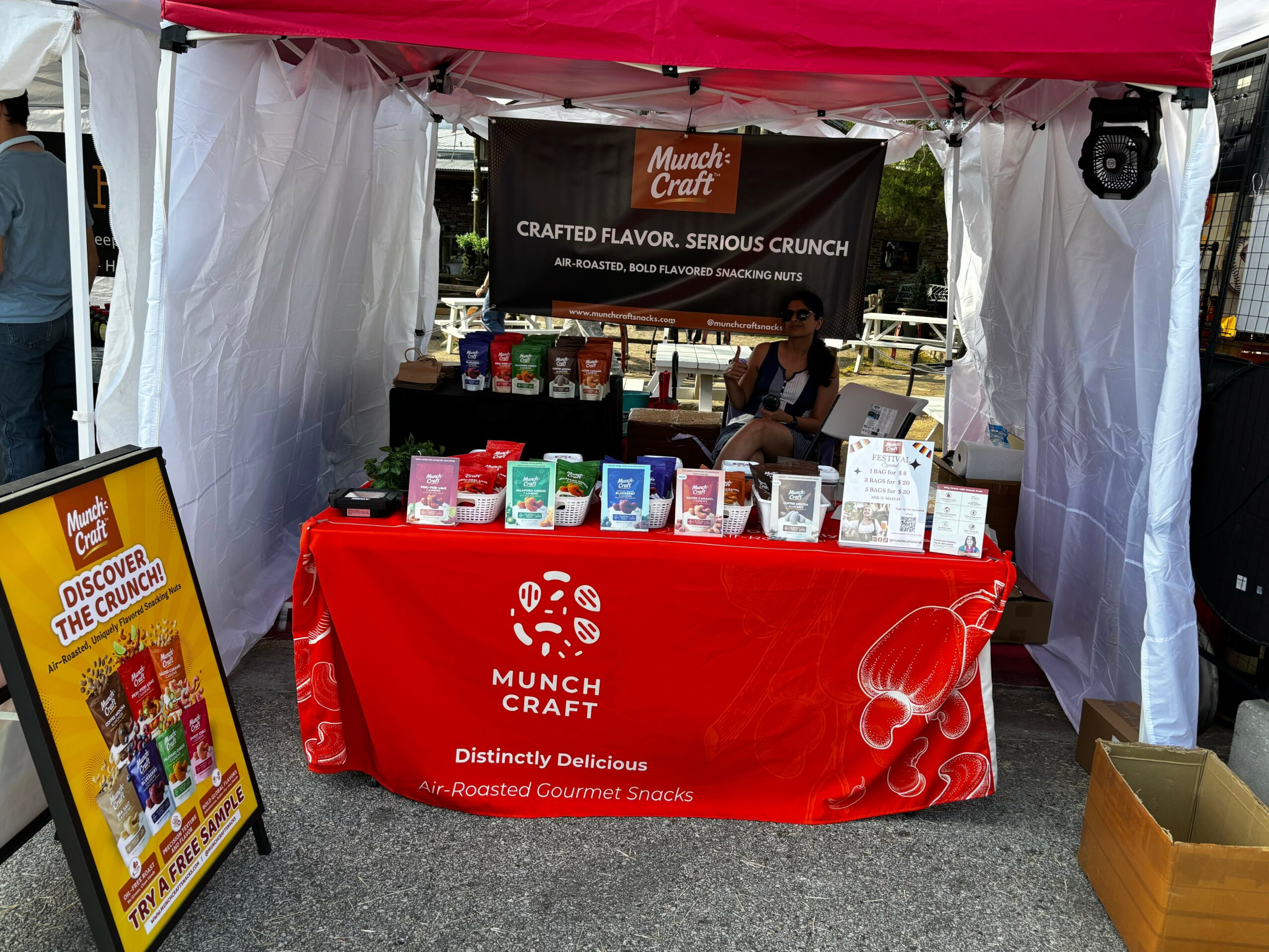 Market stall with a red tablecloth displaying snacks and drinks, under a red canopy, with a woman behind the table.