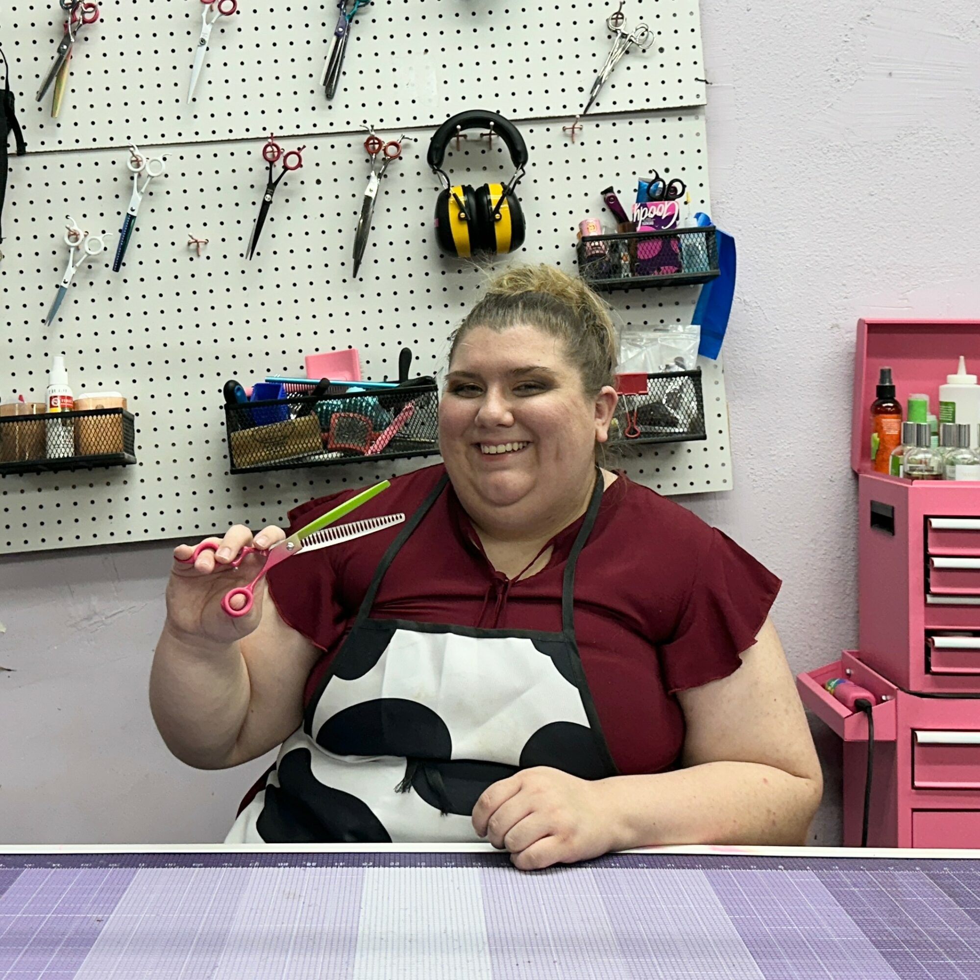 Smiling person with short hair wearing an apron, holding a pair of scissors, in a workshop with tools and supplies.