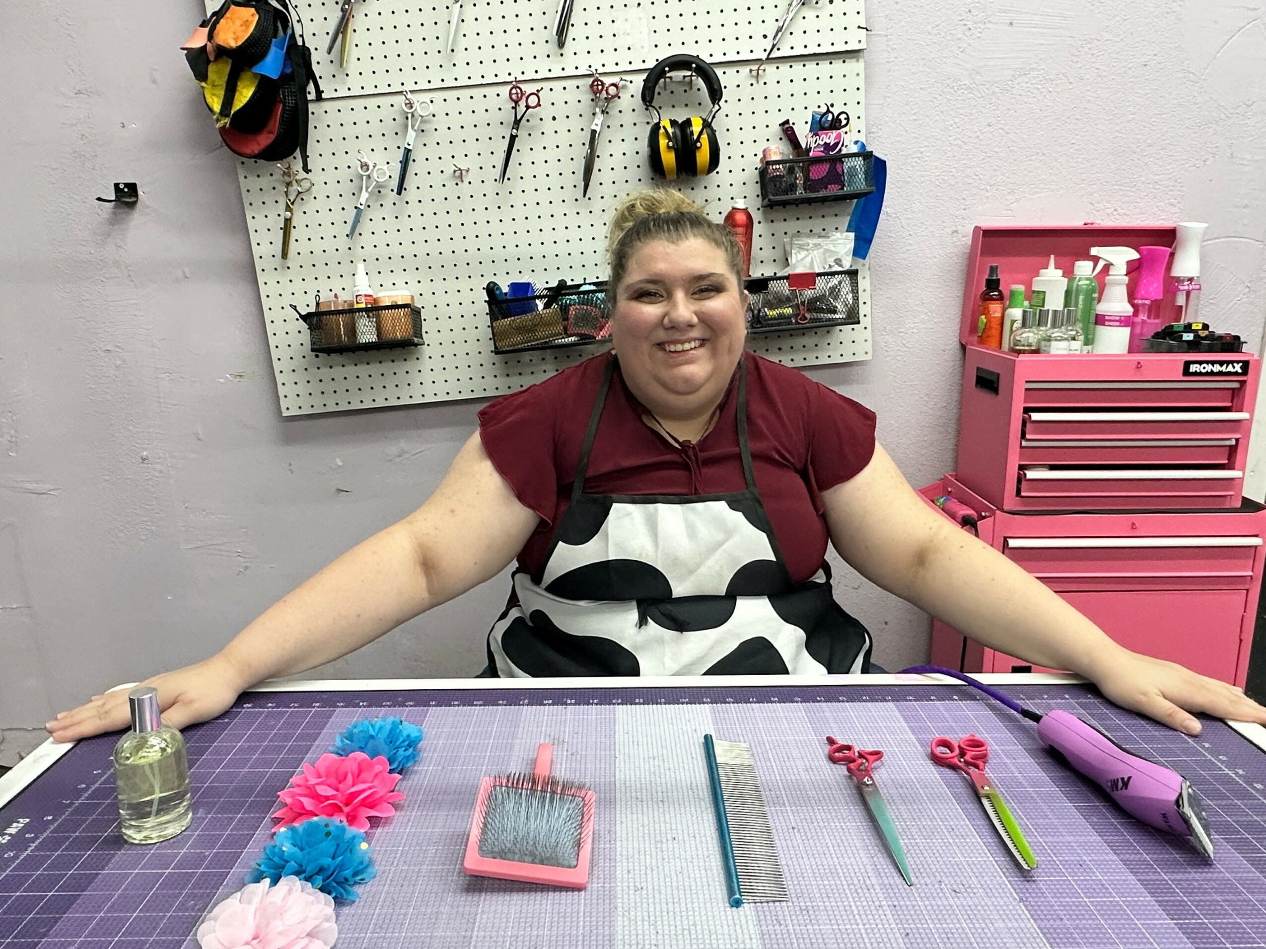 Smiling woman sitting at a work table with craft tools and supplies, workshop background with pegboard and pink storage unit.