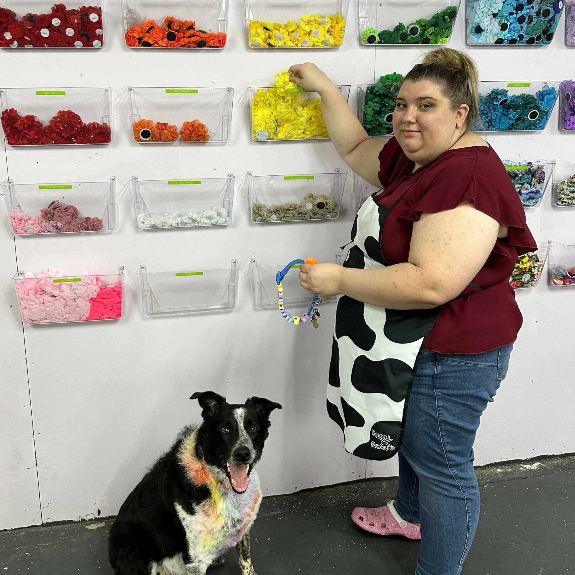 Woman selecting yellow beads from wall of colorful beads, dog sitting nearby, store setting, casual clothing, wall with organized bead containers.