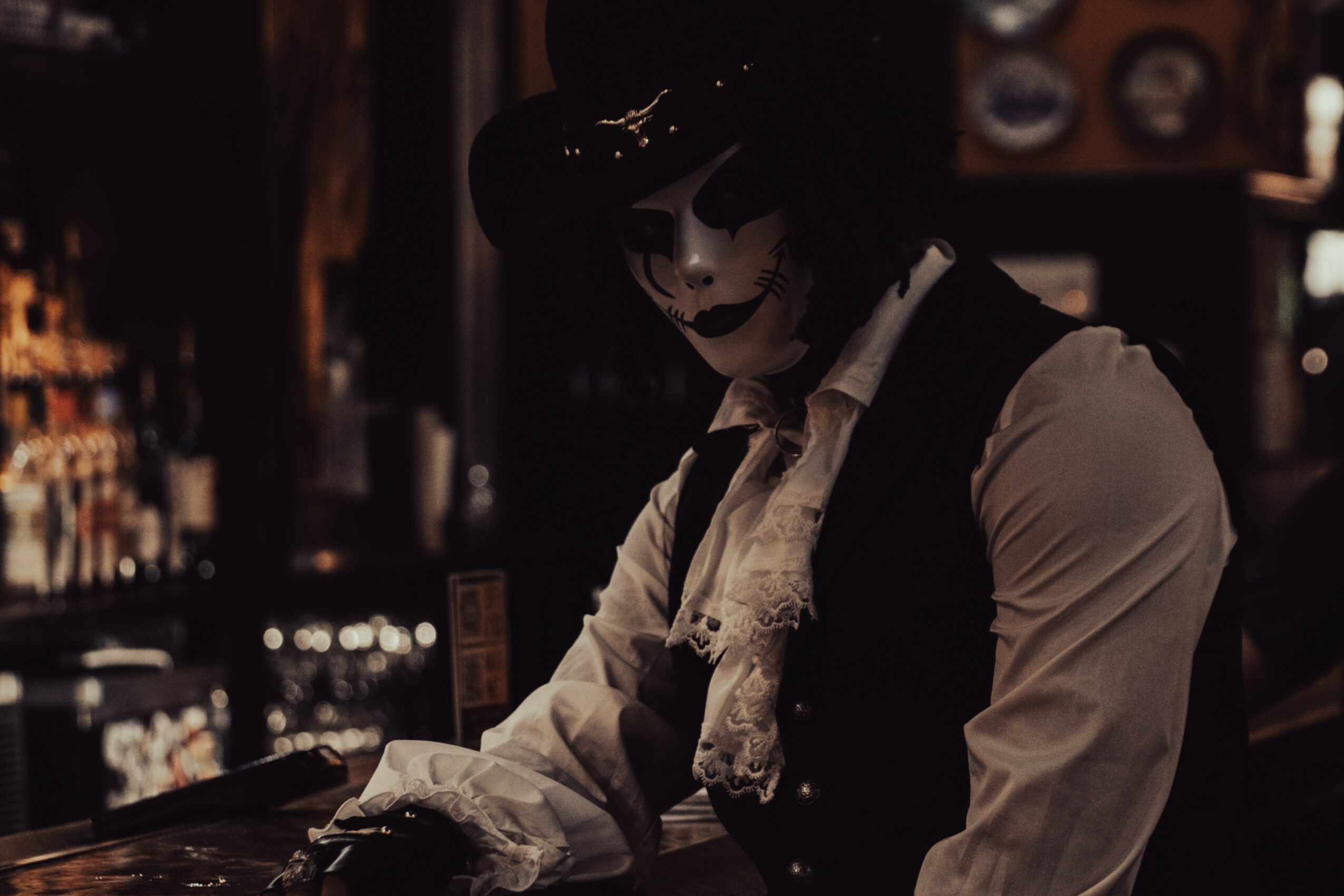 Person dressed as a mime with dark makeup, wearing a hat and vintage clothing, sitting at a bar counter.
