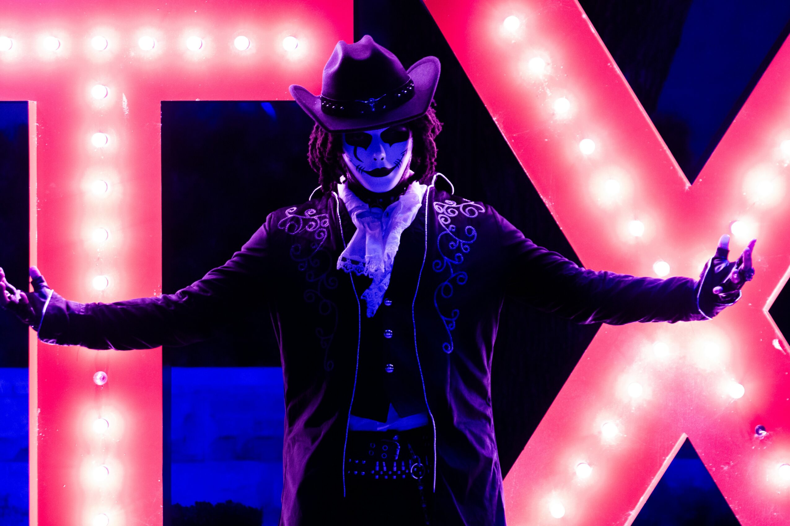 Person dressed as a skeleton in a cowboy hat with arms outstretched, illuminated by bright pink and blue lights.