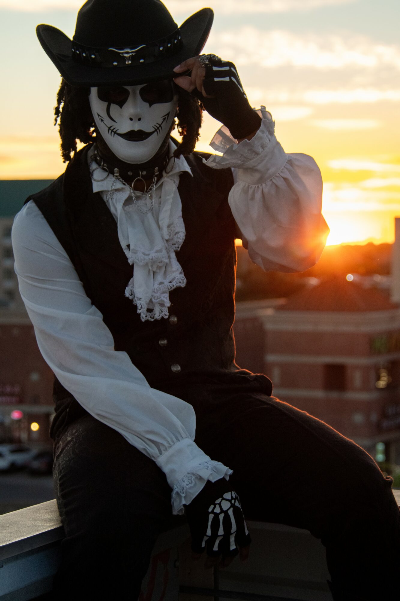 Person wearing a skeleton mask, black hat, and white blouse, sitting outdoors during sunset.