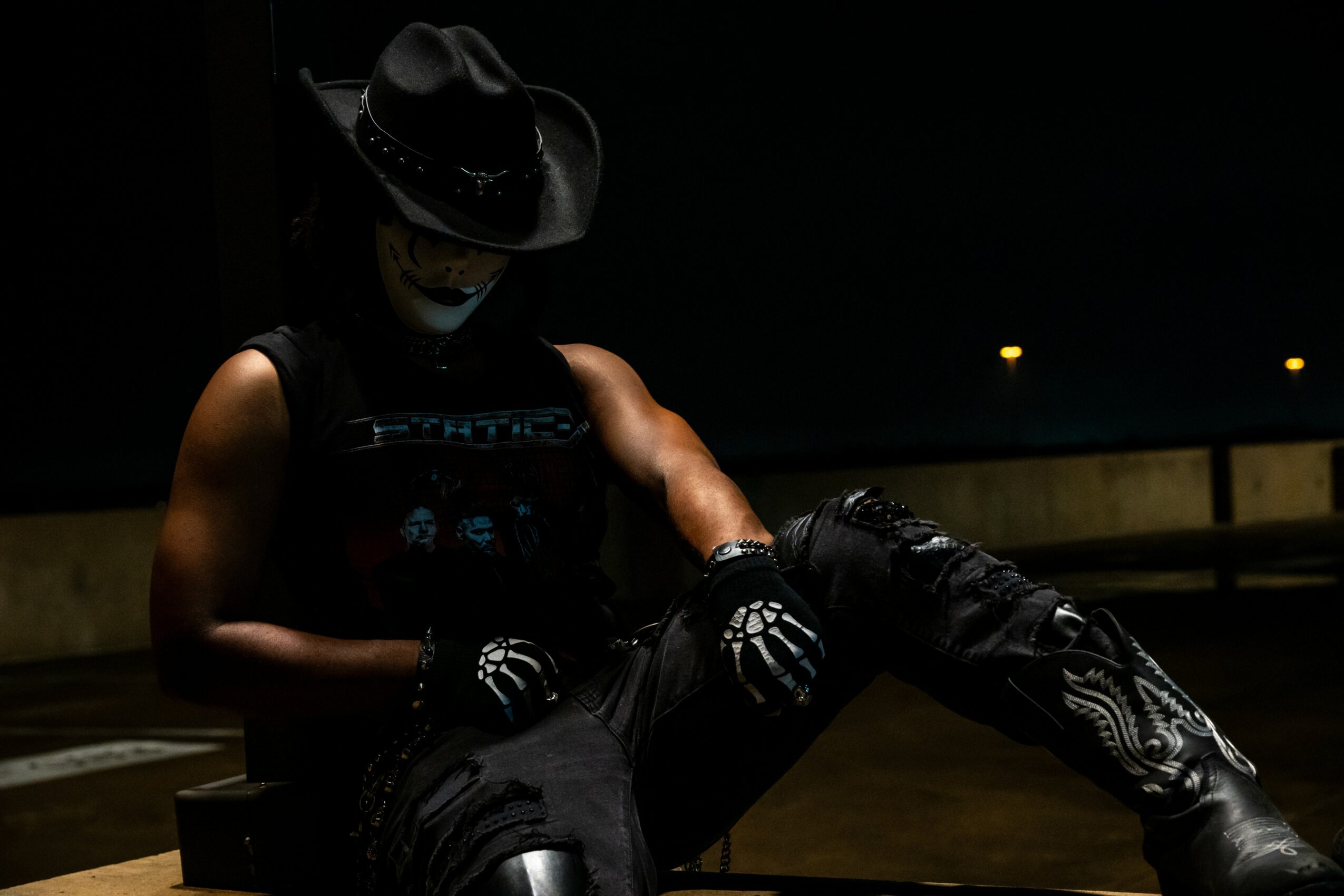 Person wearing a hat, mask, and gloves sitting on a motorcycle at night, with a dark background and streetlights.