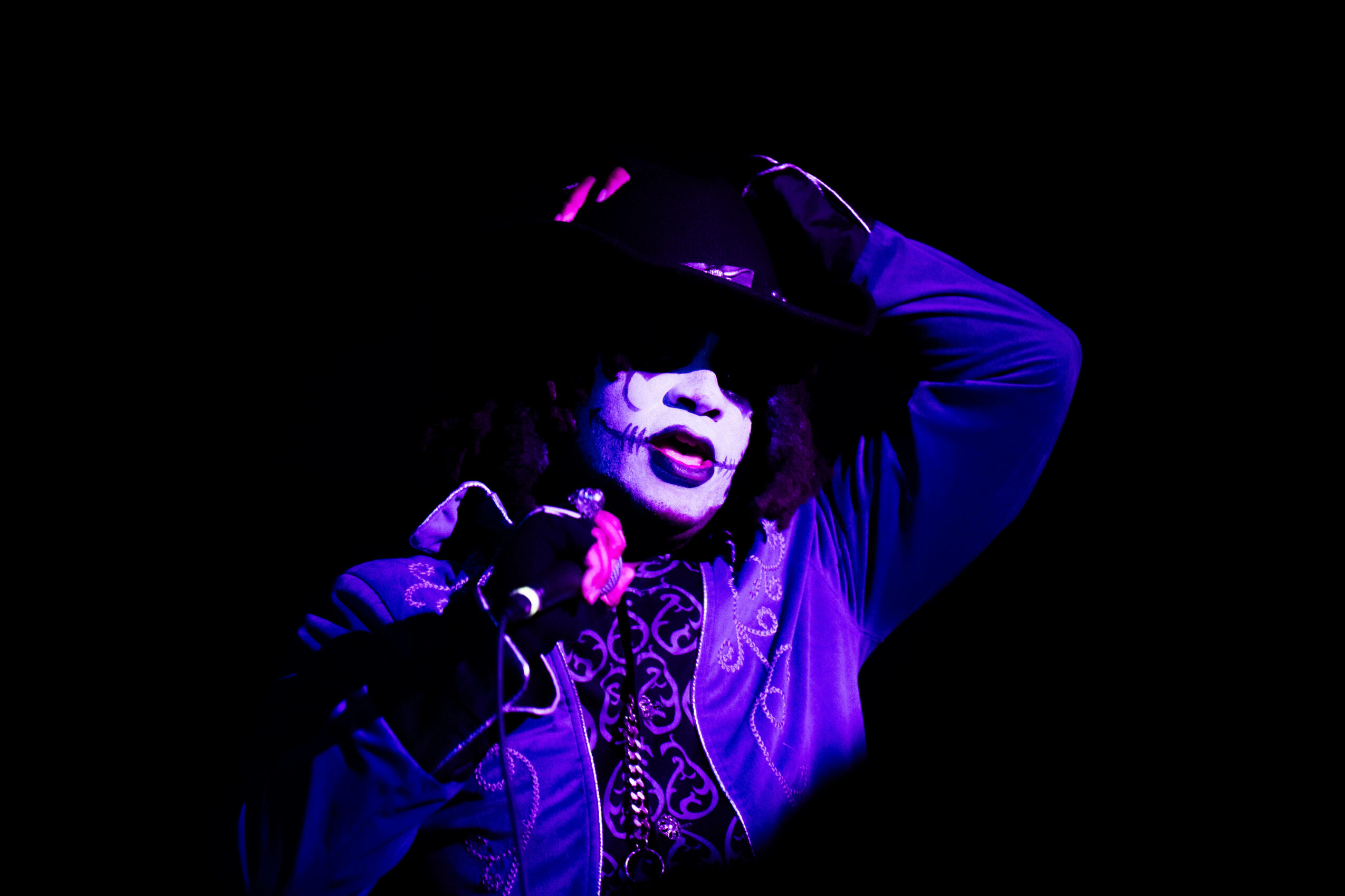 Performer with face painted, wearing a hat and purple costume, singing into a microphone in a dark setting.