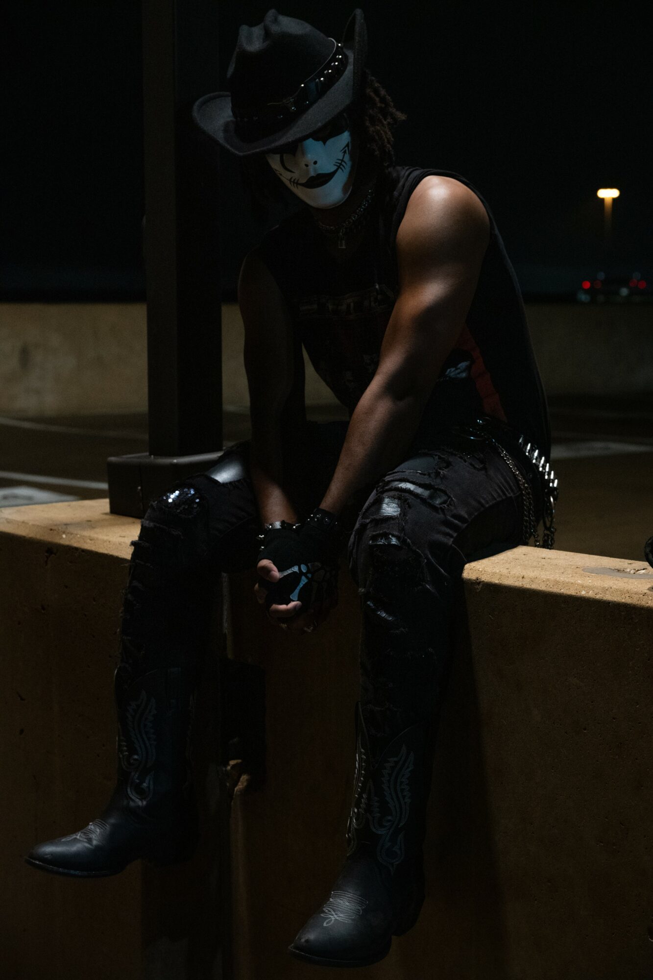 Person dressed in dark clothing with face painted white, sitting on a ledge at night, wearing a hat and boots.