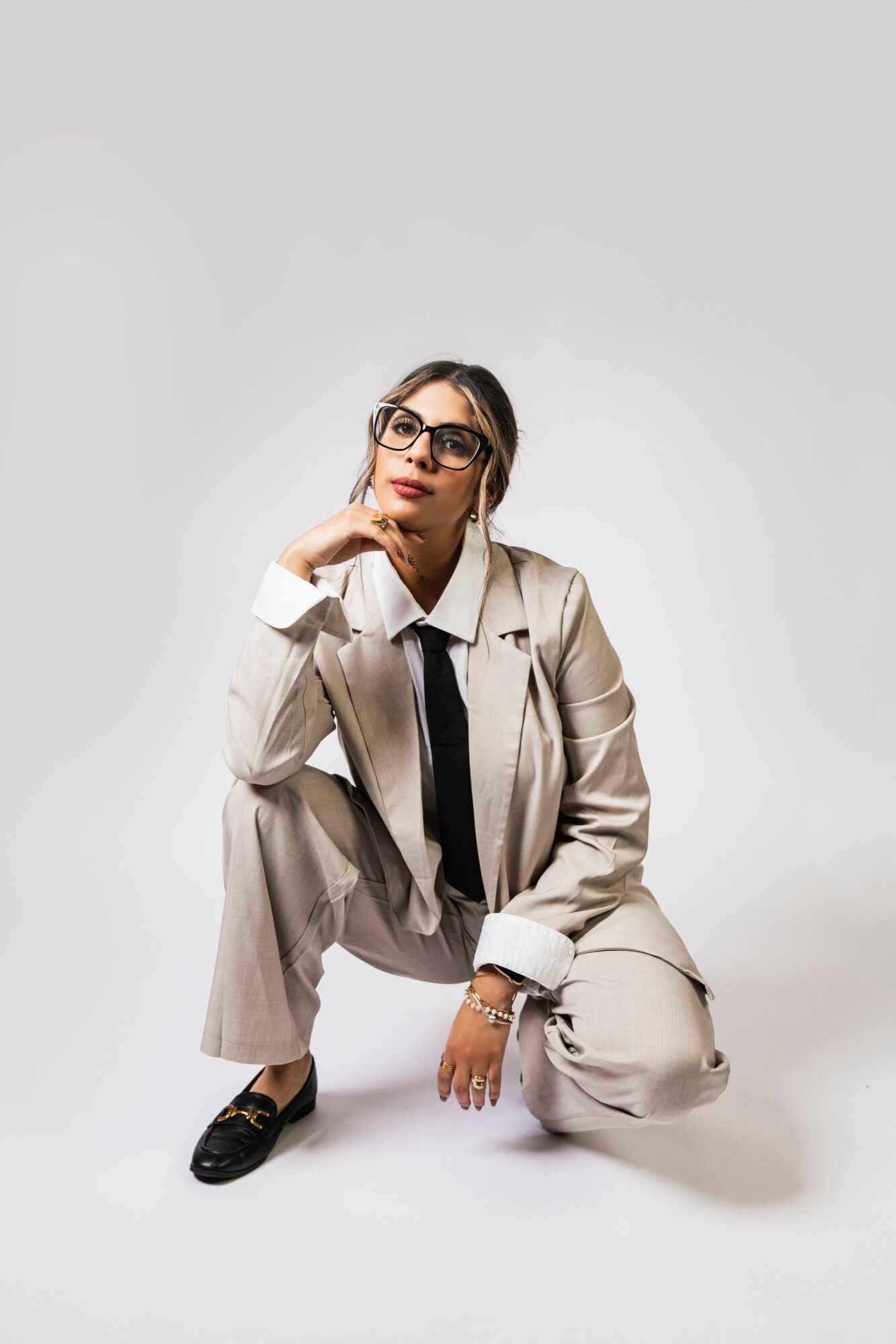 Woman in beige suit crouching, wearing glasses, with hand on chin, against plain light background.