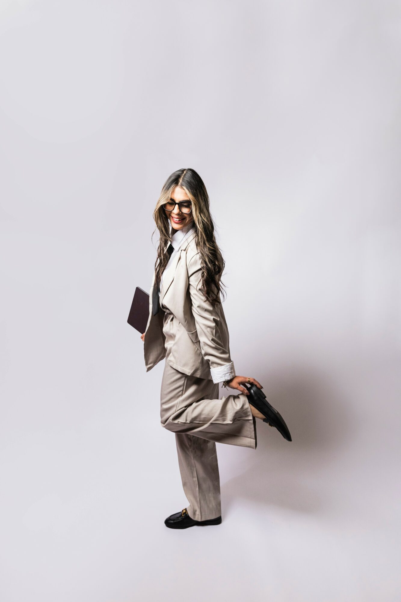 Woman in business attire holding a tablet and a shoe, smiling, standing against a plain background.