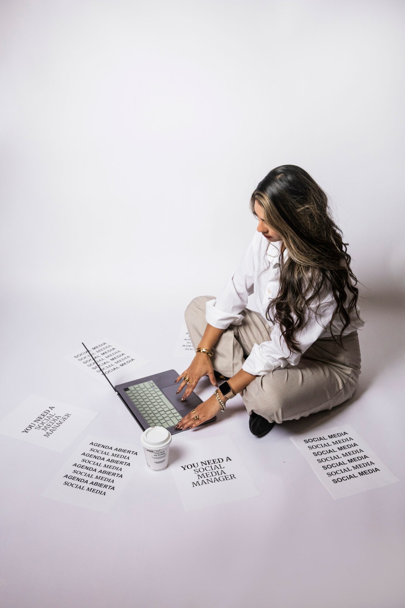 Woman sitting on the floor with a laptop, surrounded by papers and a cup, against a plain white background.