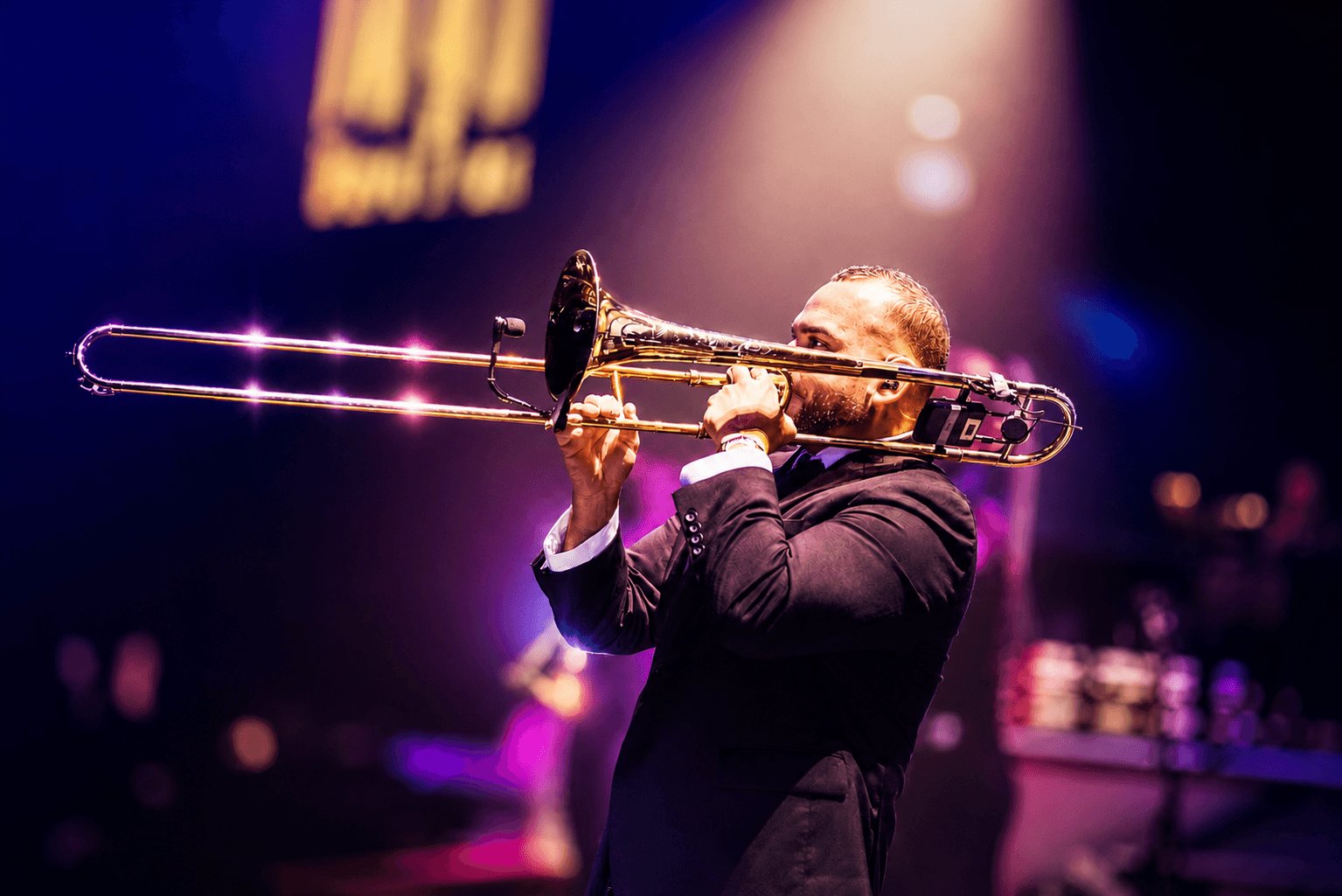 Person playing trombone on stage with colorful lighting, wearing a dark suit, in a concert setting.