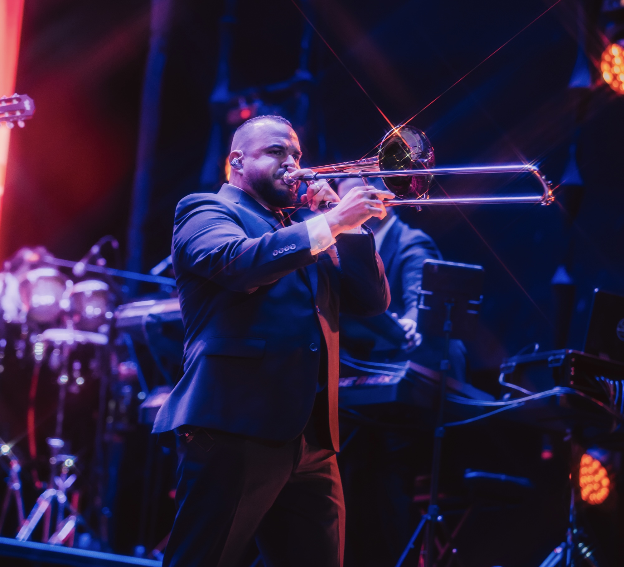Man playing trombone on stage with colorful lighting and musical instruments in background.