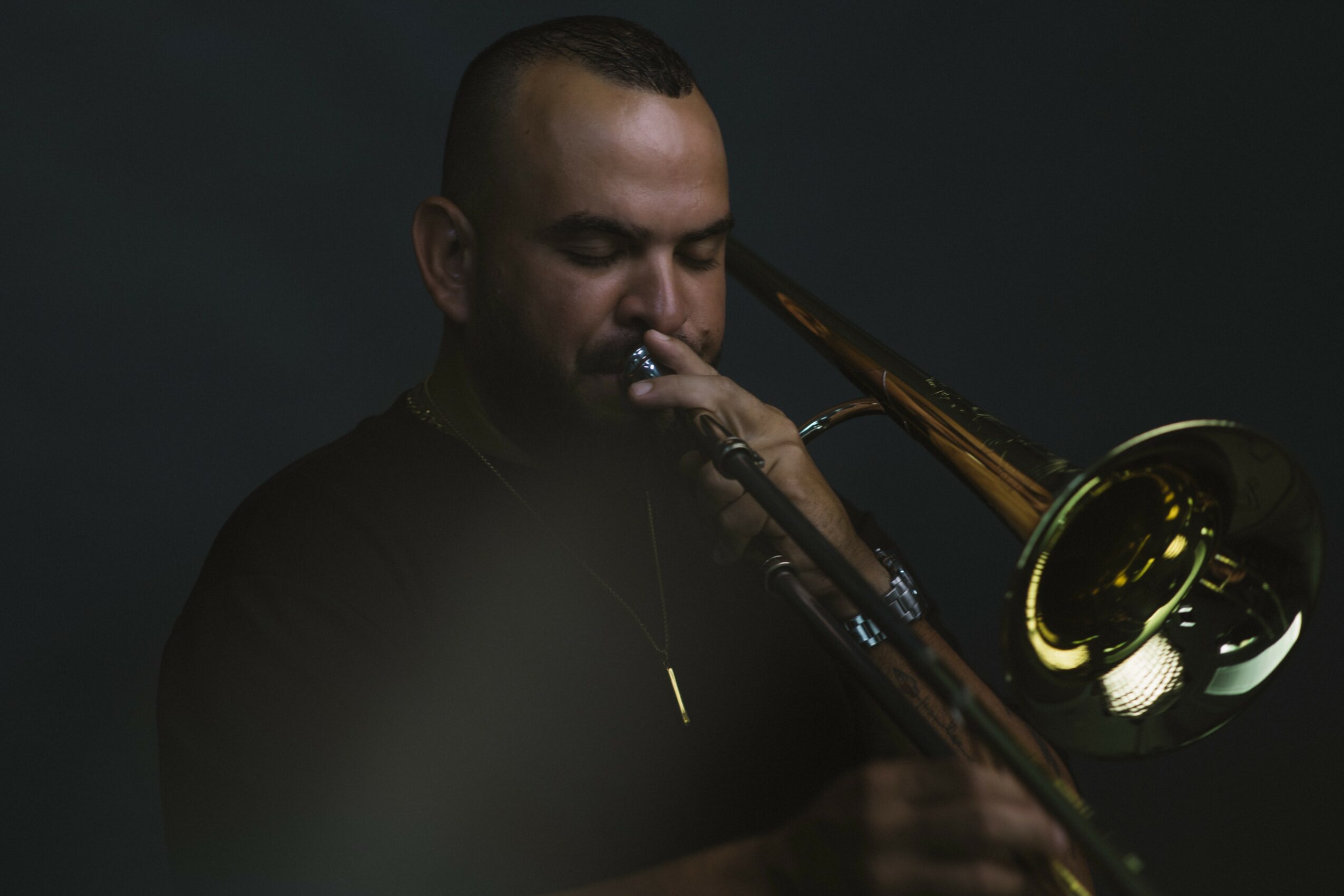 Person playing a trombone with a dark background, eyes closed, focused on music, wearing dark clothing.