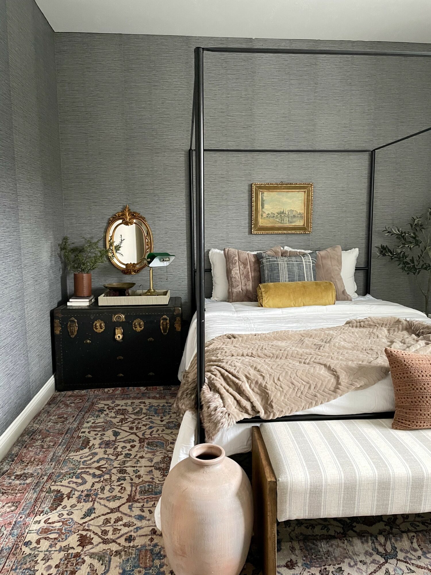 Bedroom with gray textured wall, bed with pillows and beige blanket, black nightstand, decorative mirror, framed picture, and striped bench.