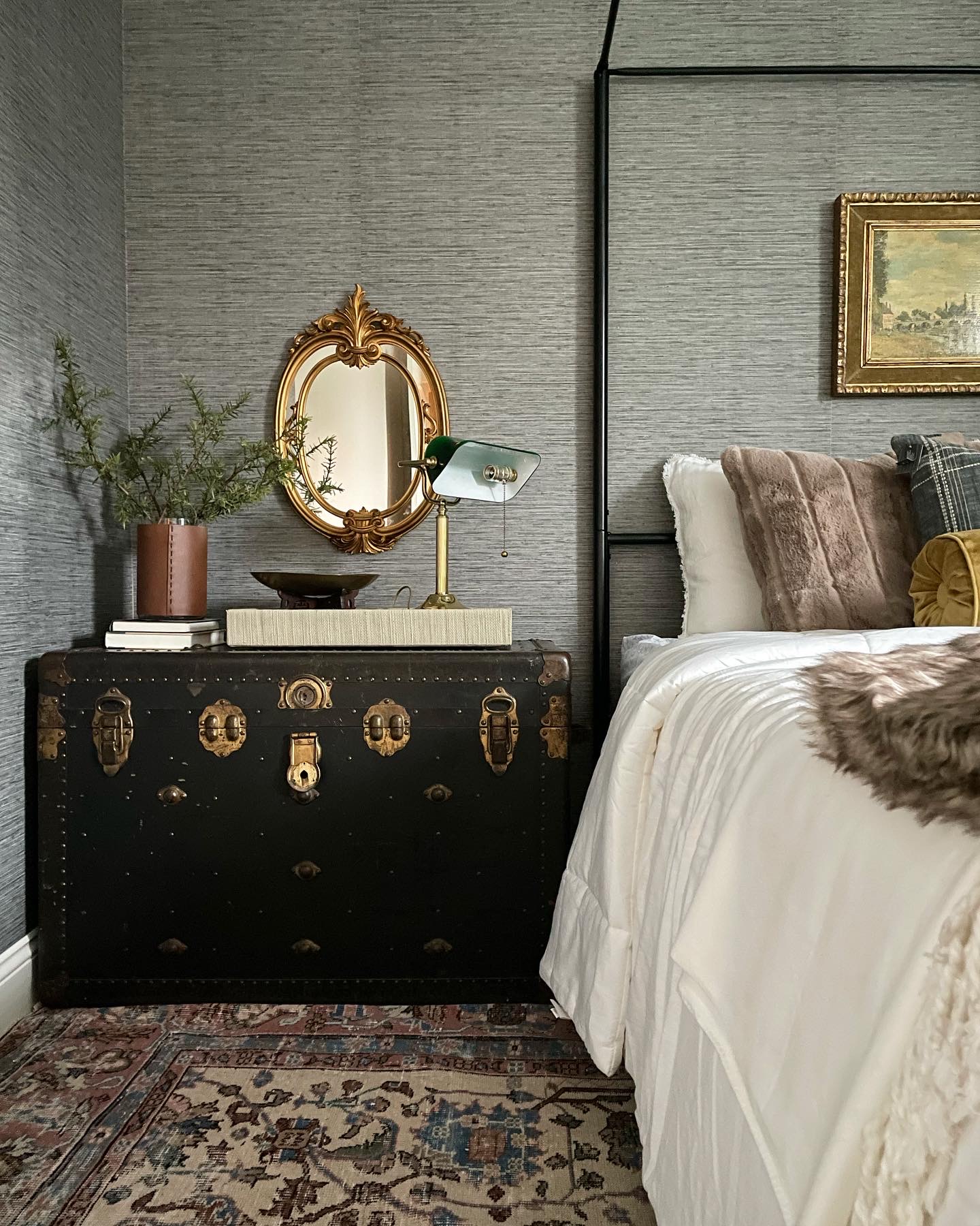 Bedroom corner with a black chest, gold mirror, table lamp, and a bed with pillows and a blanket.