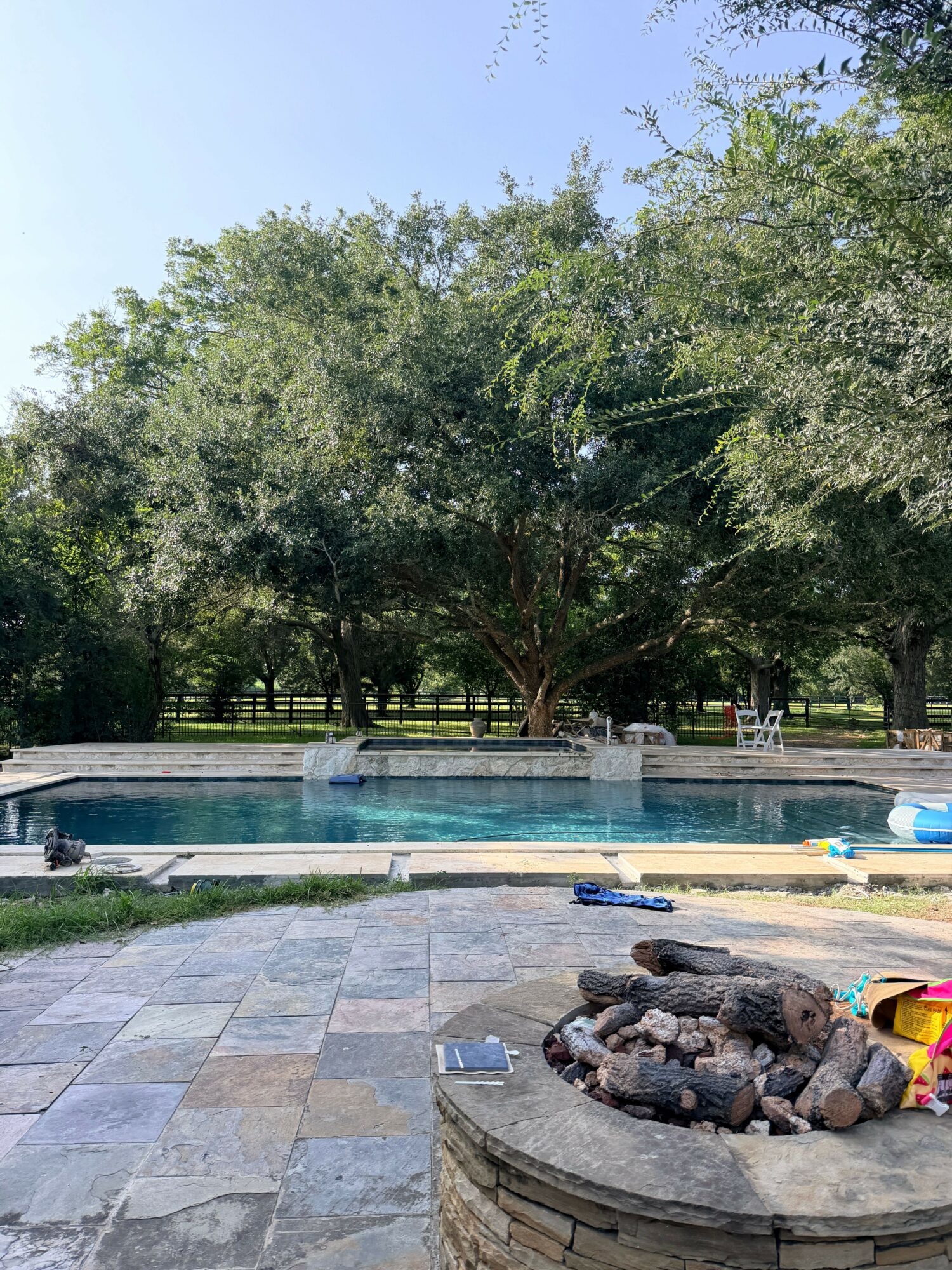 Swimming pool with trees and a fire pit nearby, outdoor patio area with toys and chairs, sunny day.