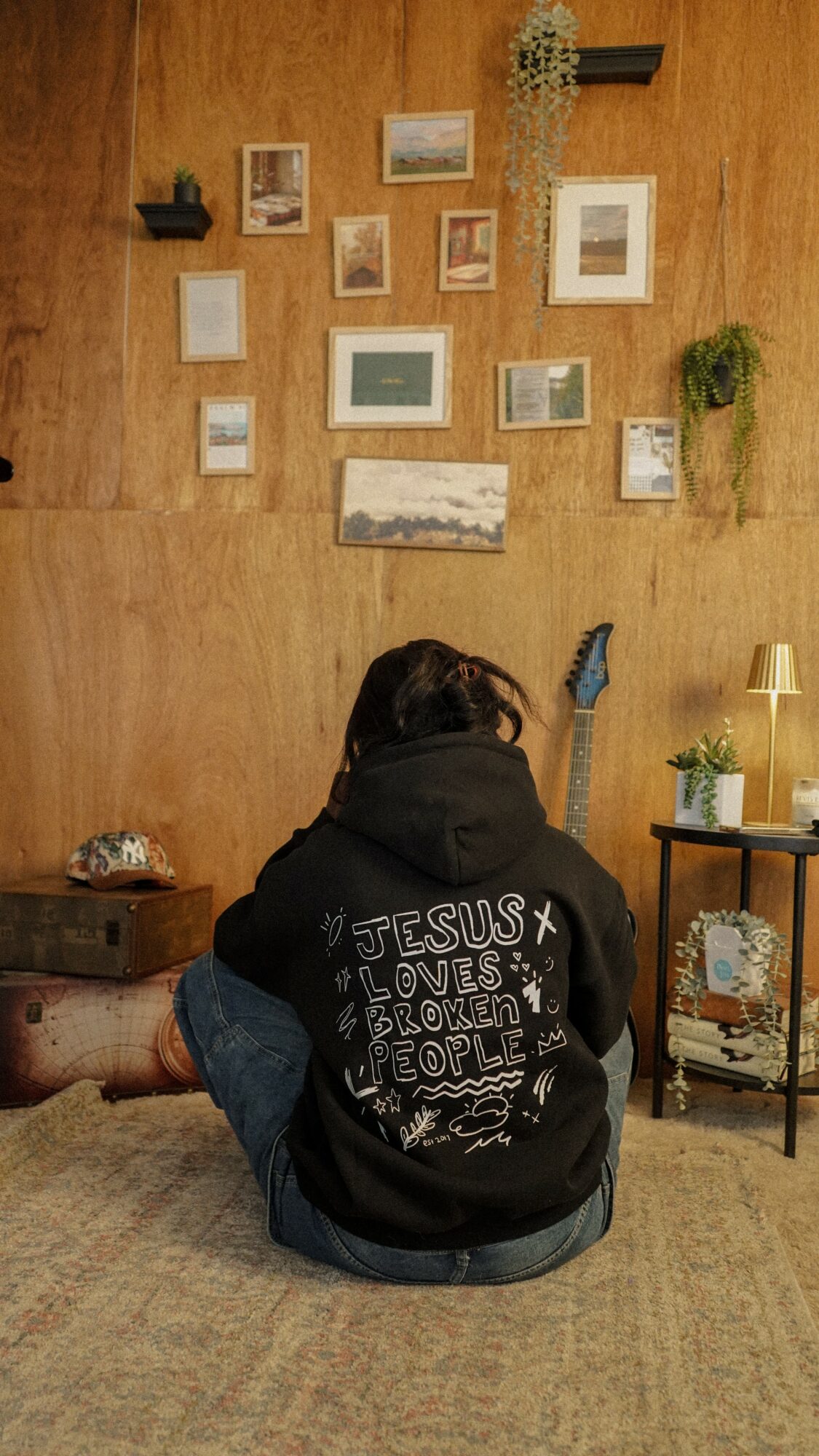 Person sitting on the floor with back to camera, wearing a black hoodie with white text, in a cozy room with wall art.