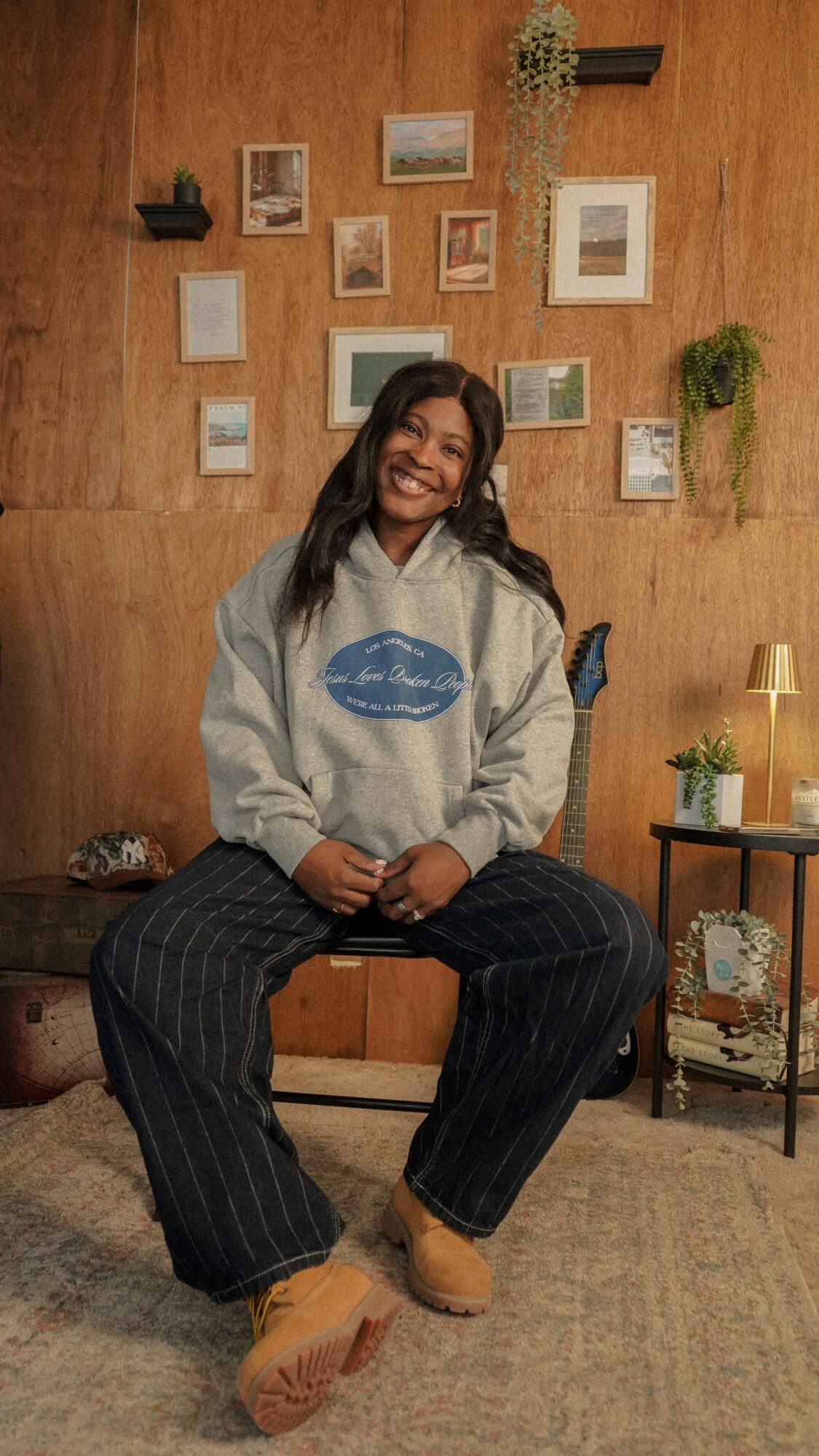 Person sitting on a chair smiling, wearing a hoodie and striped pants, in a cozy room with wall art and plants