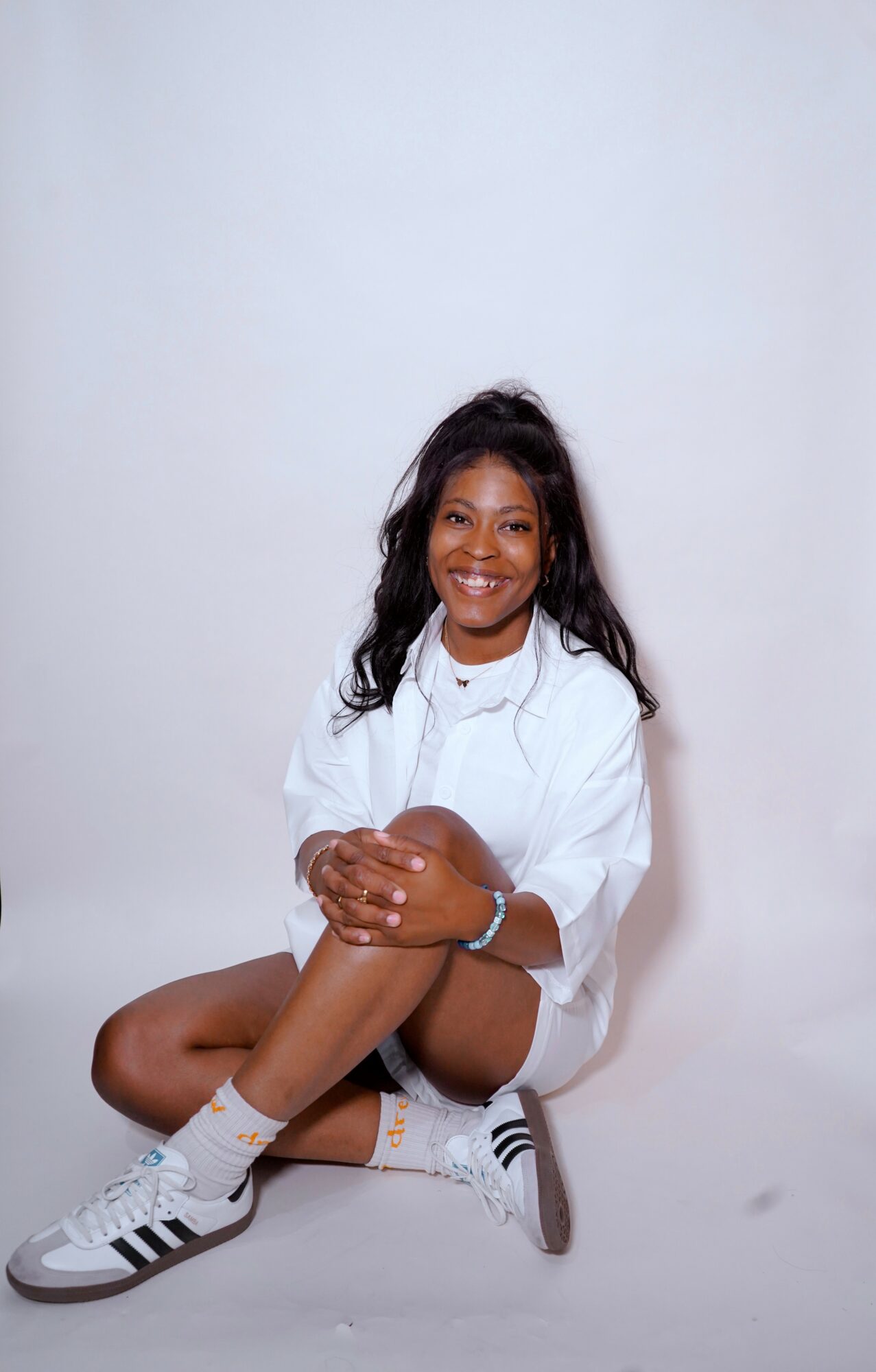 Young woman sitting on the floor, smiling, wearing a white shirt, shorts, and sneakers, against a plain background.