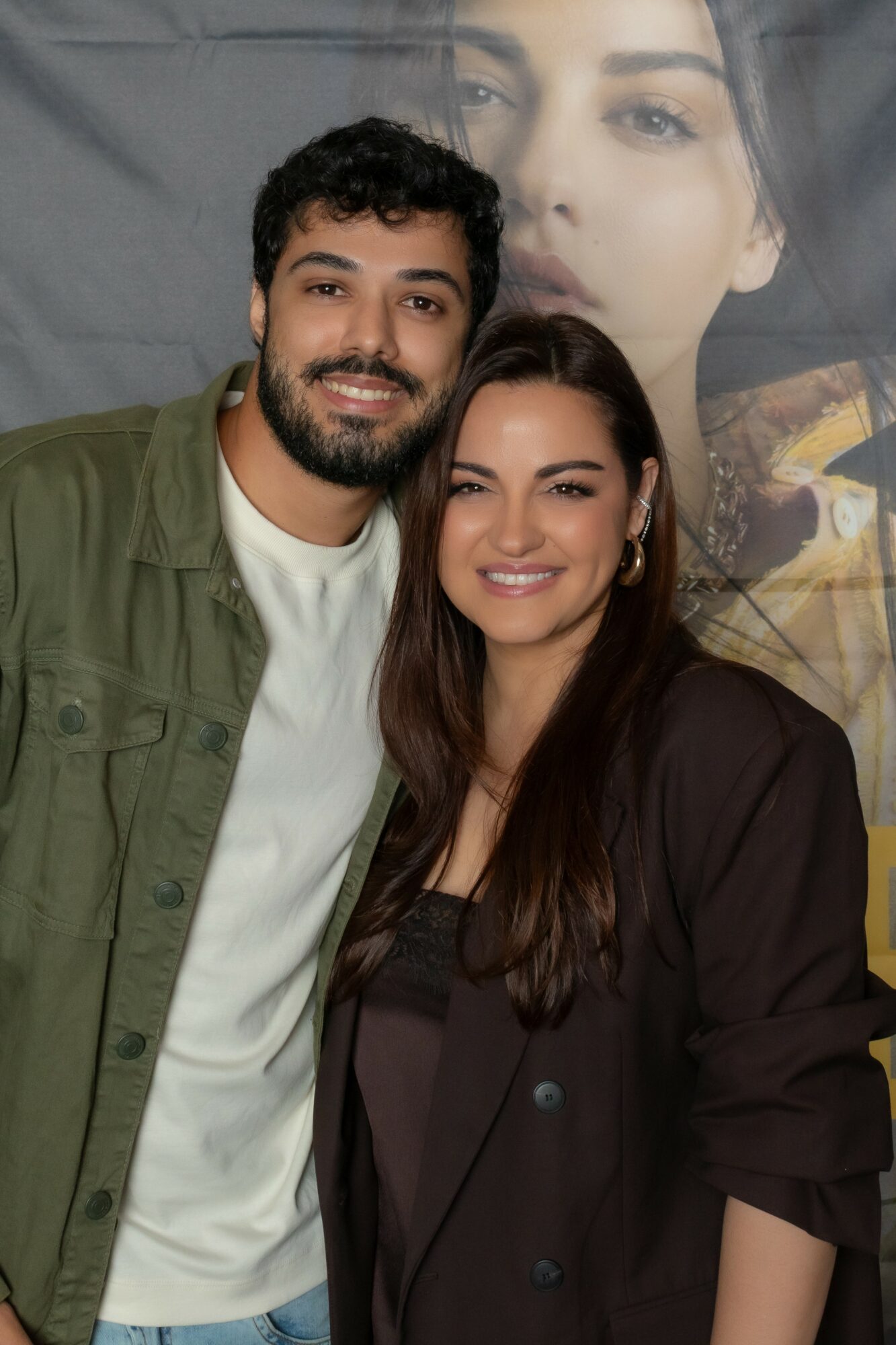 Two people smiling, standing close together, with a large portrait of a woman's face in the background.