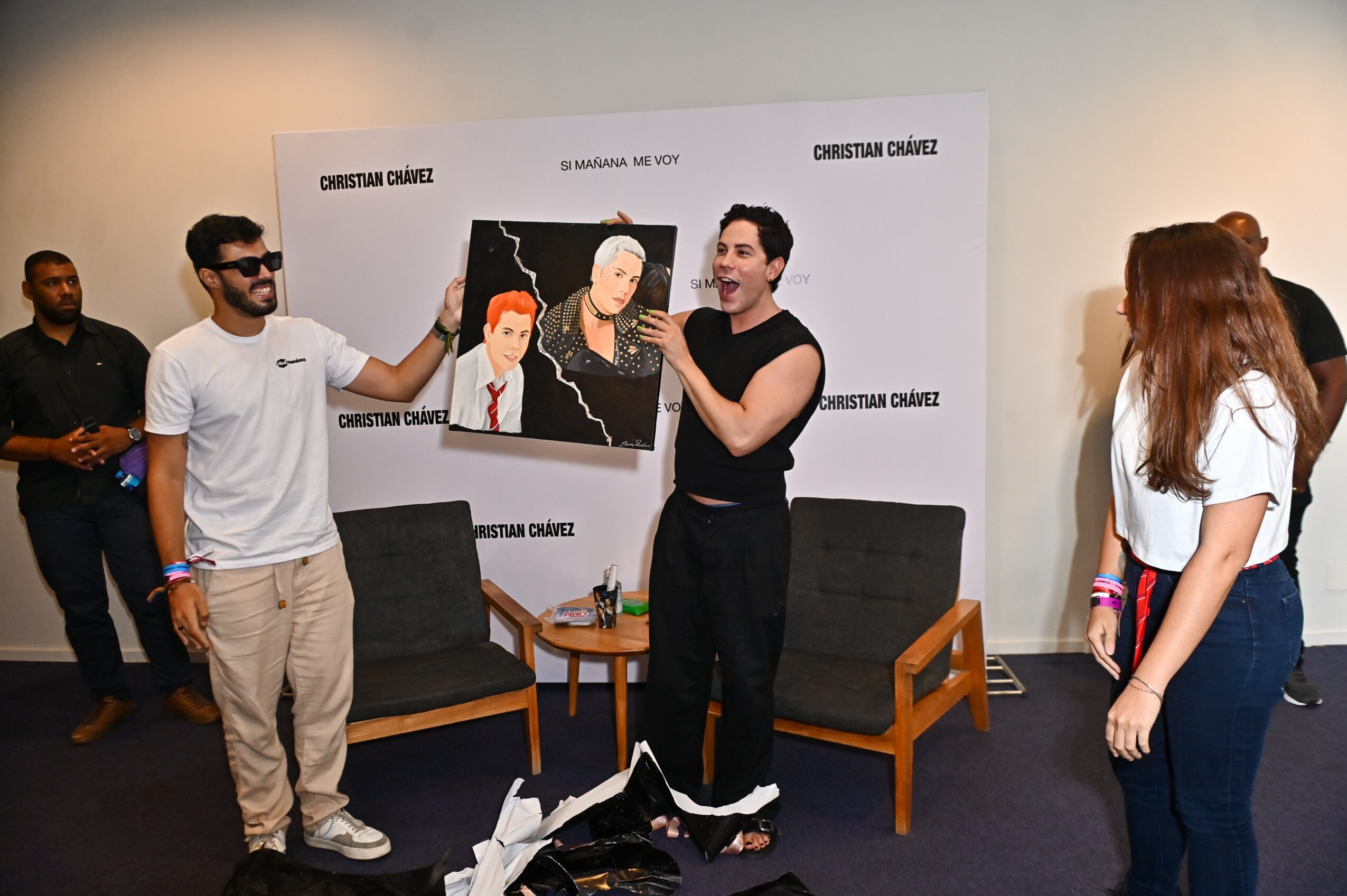 Group of people in a room, one person holding a painting, others watching, with chairs and a table, background with text.