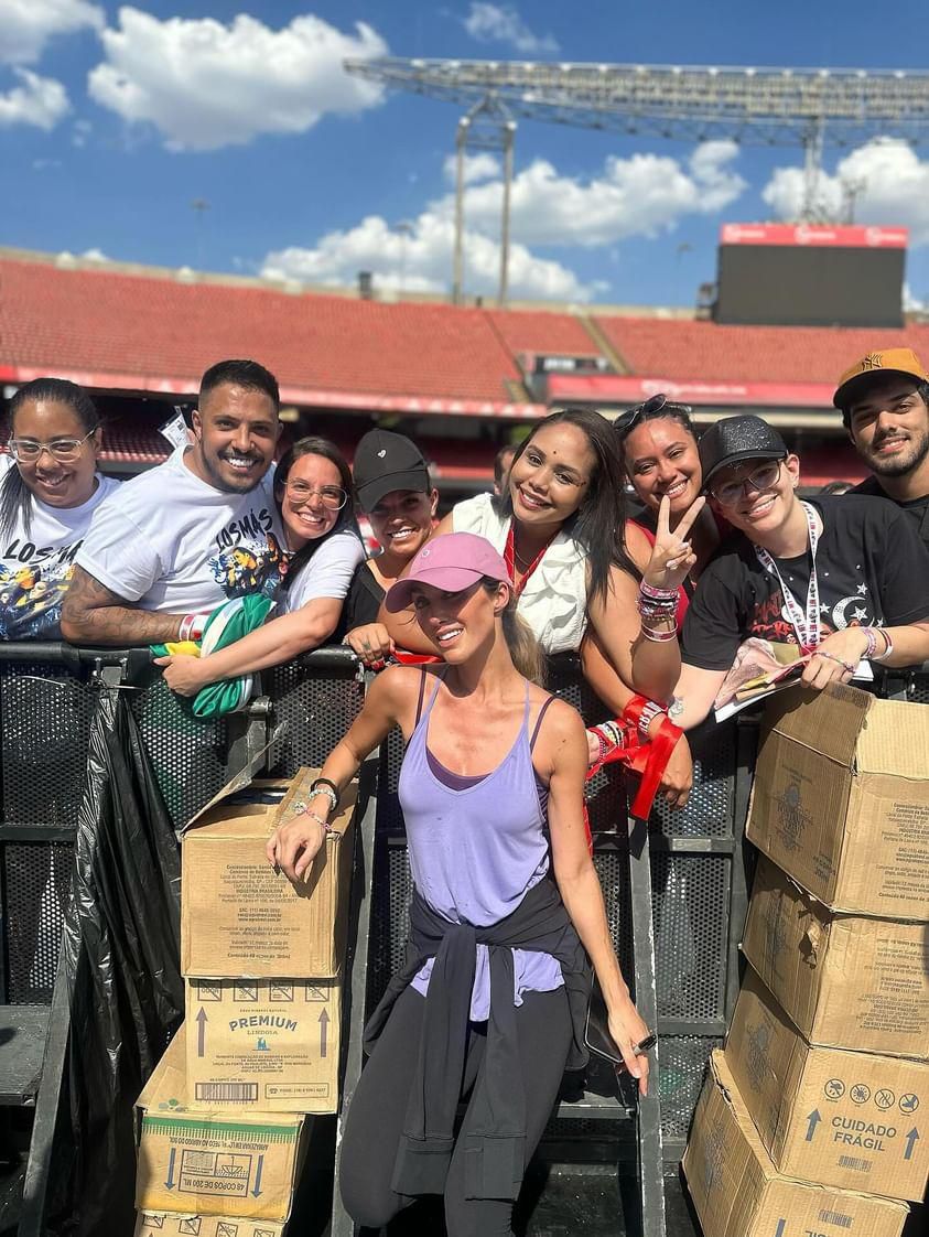 Group of people smiling outdoors, some making peace signs, with boxes and a stadium in the background.