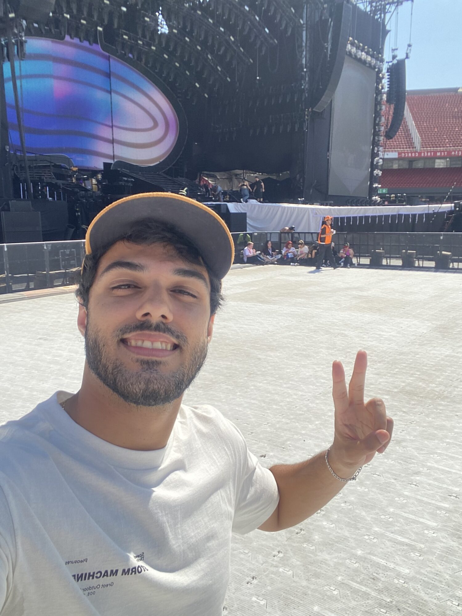 Young man with a beard and cap smiling and making a peace sign outdoors, stage and buildings in background.