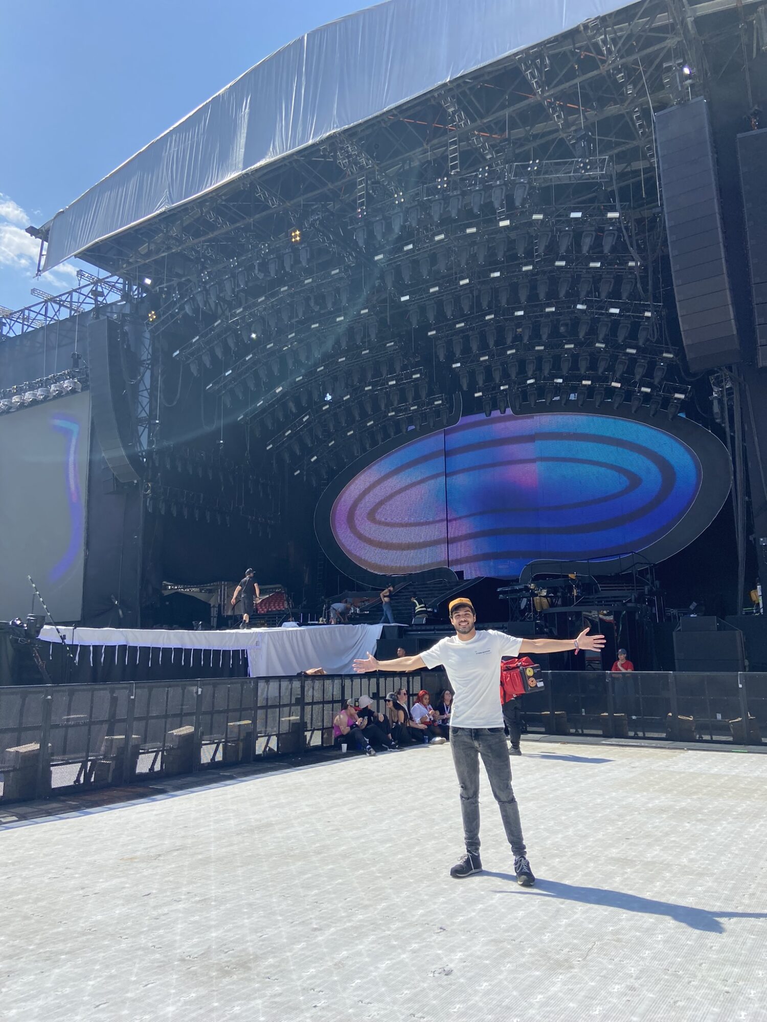 Person standing with arms outstretched in front of a large outdoor stage with a colorful circular light display, blue sky above.