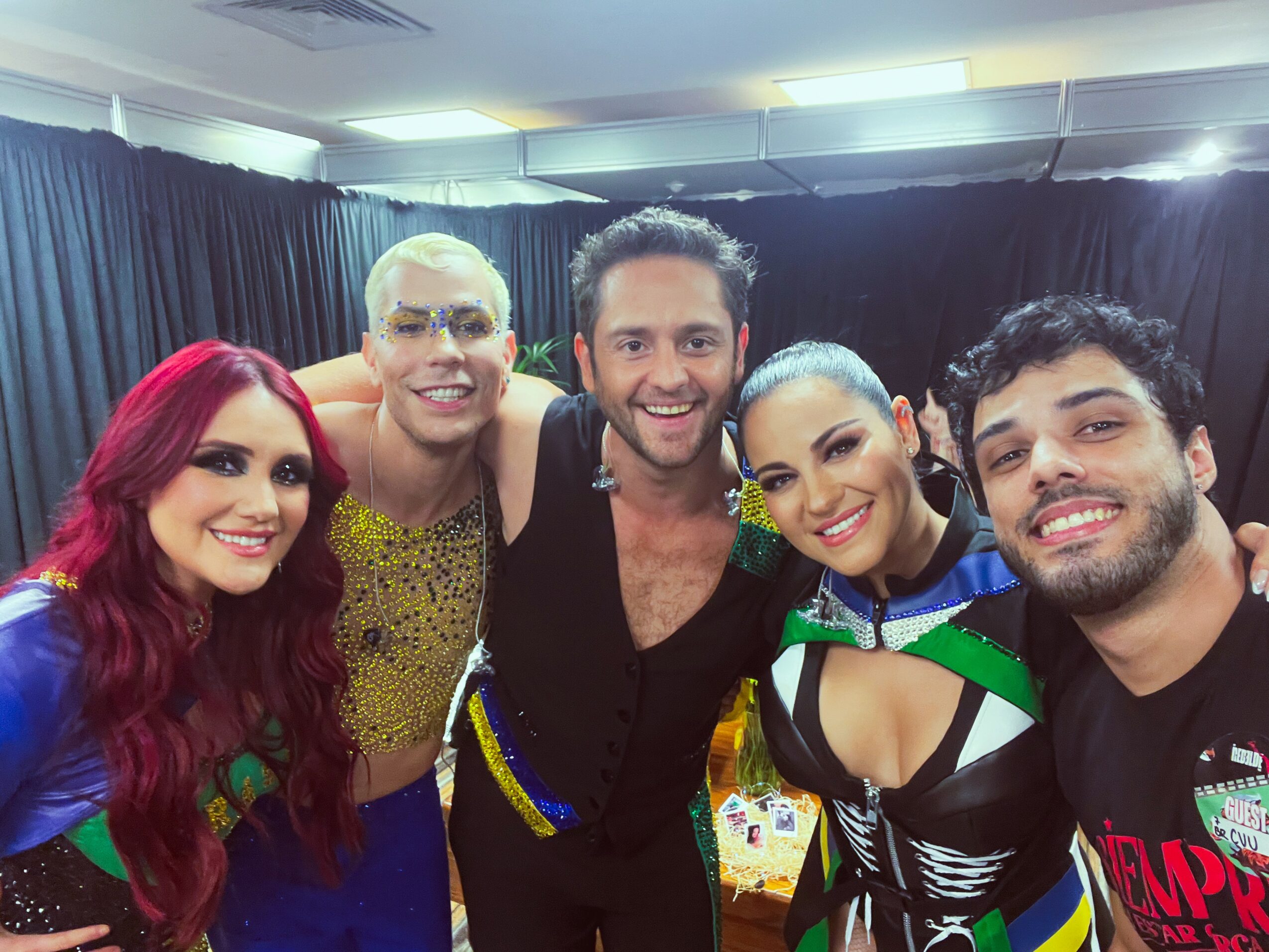 Group of five people smiling, dressed in colorful costumes, standing indoors with dark curtains behind them.