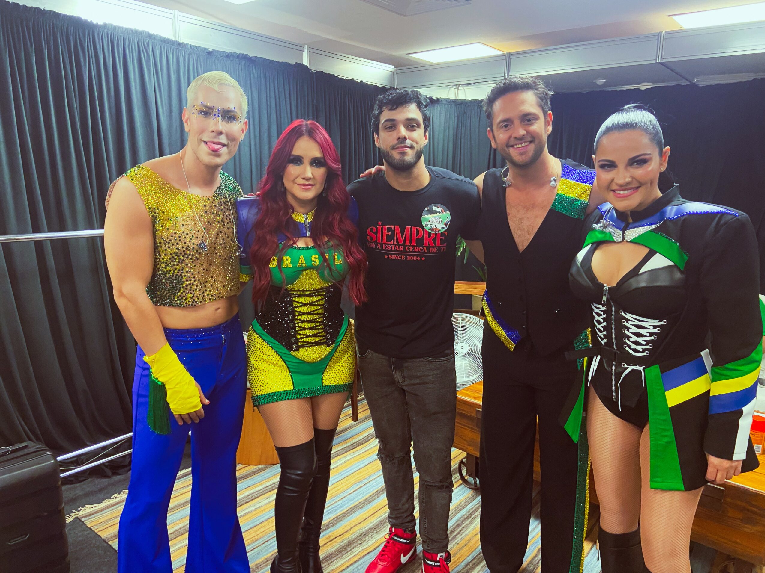 Five people standing together, four women in colorful costumes and one man in casual clothing, smiling indoors.