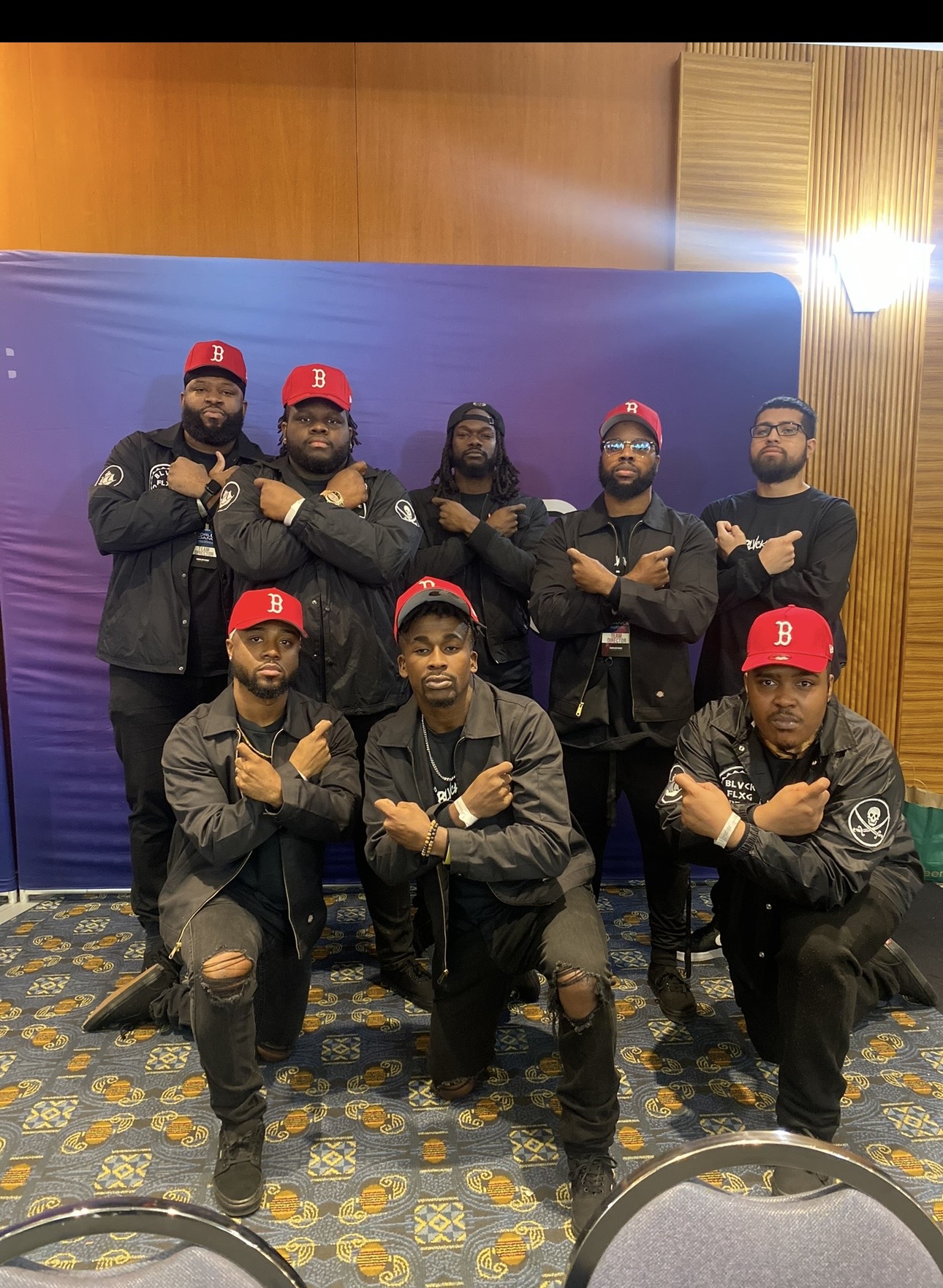 Group of nine men posing indoors, some kneeling and some standing, wearing black jackets and red caps, with a blue backdrop behind them.