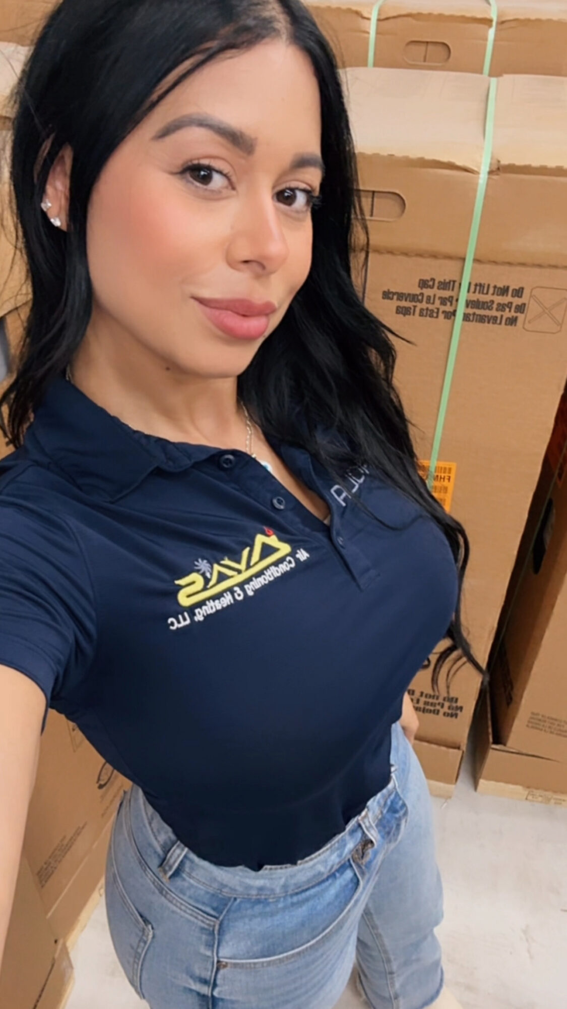 Woman with dark hair wearing a navy polo shirt and light jeans, standing in front of cardboard boxes.