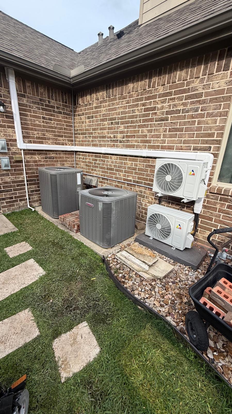 Air conditioning units and a heat pump installed outside a brick building with a grassy area and stepping stones.
