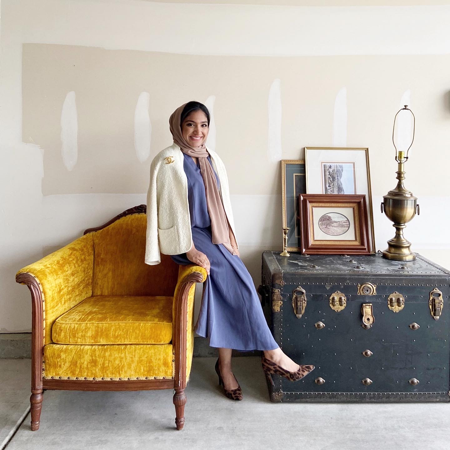 Woman sitting on a yellow velvet sofa, wearing a blue dress and beige cardigan, with a scarf on her head, in a room with vintage decor.