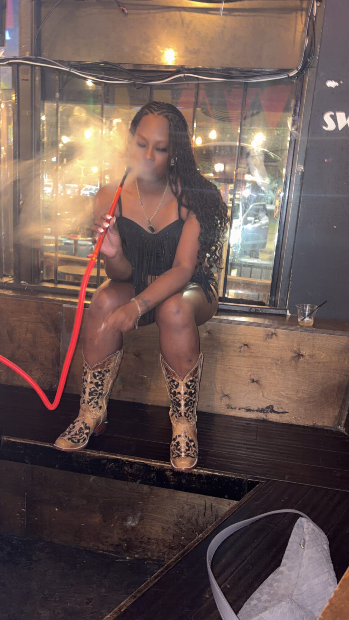 Woman sitting on a windowsill at night, with city lights outside, wearing a black dress and patterned boots.