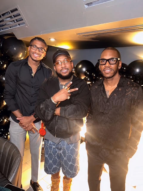 Three men standing indoors, smiling, with black balloons in the background, one making a peace sign, casual attire, warm lighting.