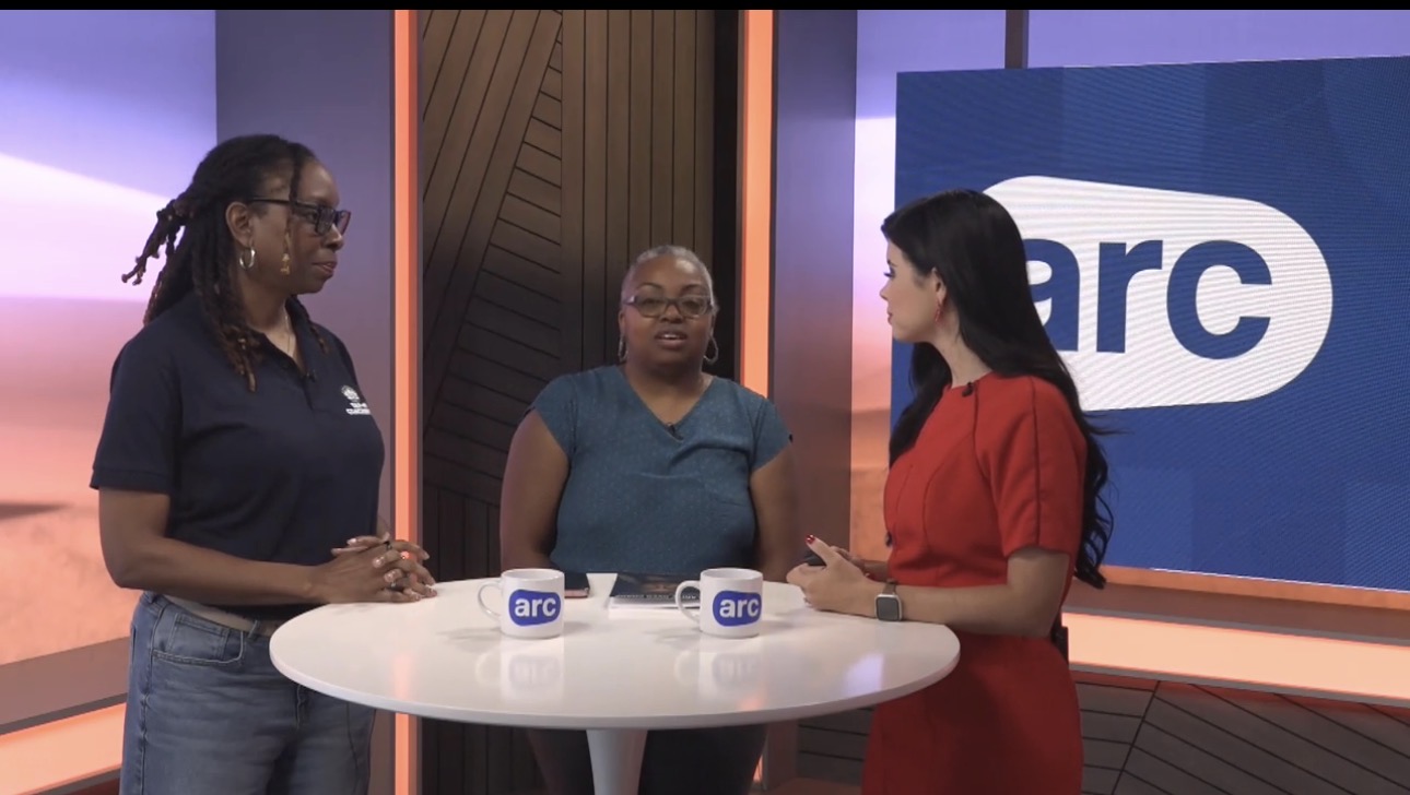 Three women standing around a table with two cups, engaged in conversation, in a studio with a large 'arc' logo background.