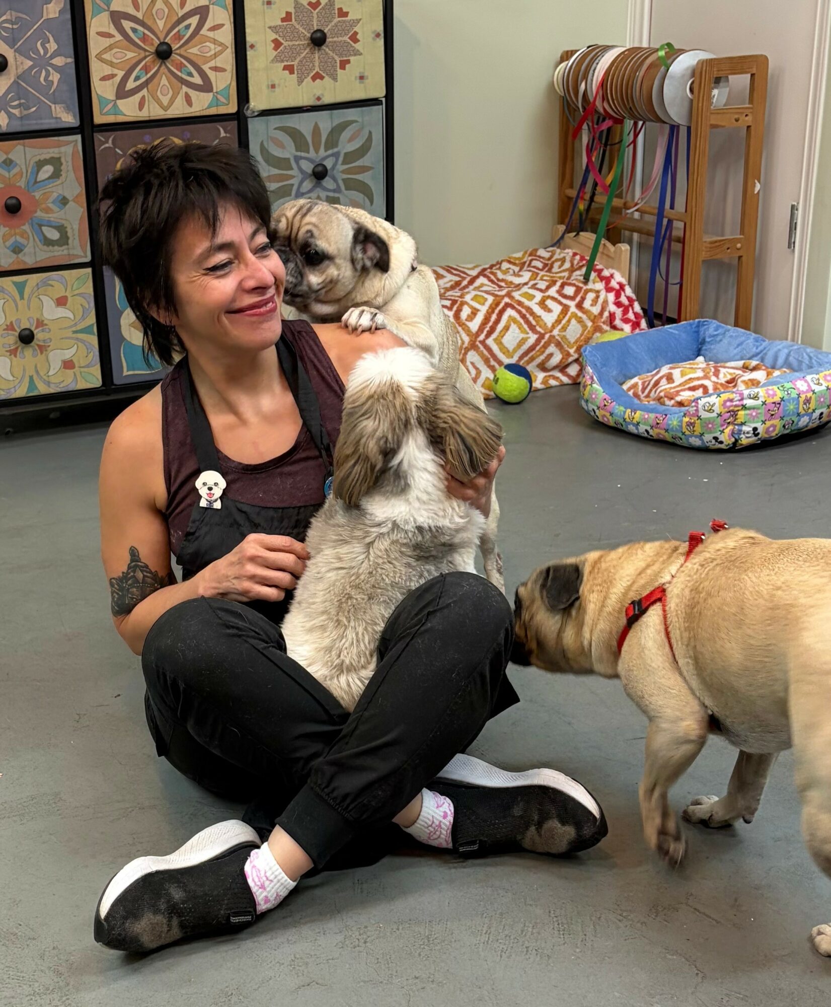 Woman sitting on the floor with three dogs, smiling, in a room with colorful tiles and pet supplies.