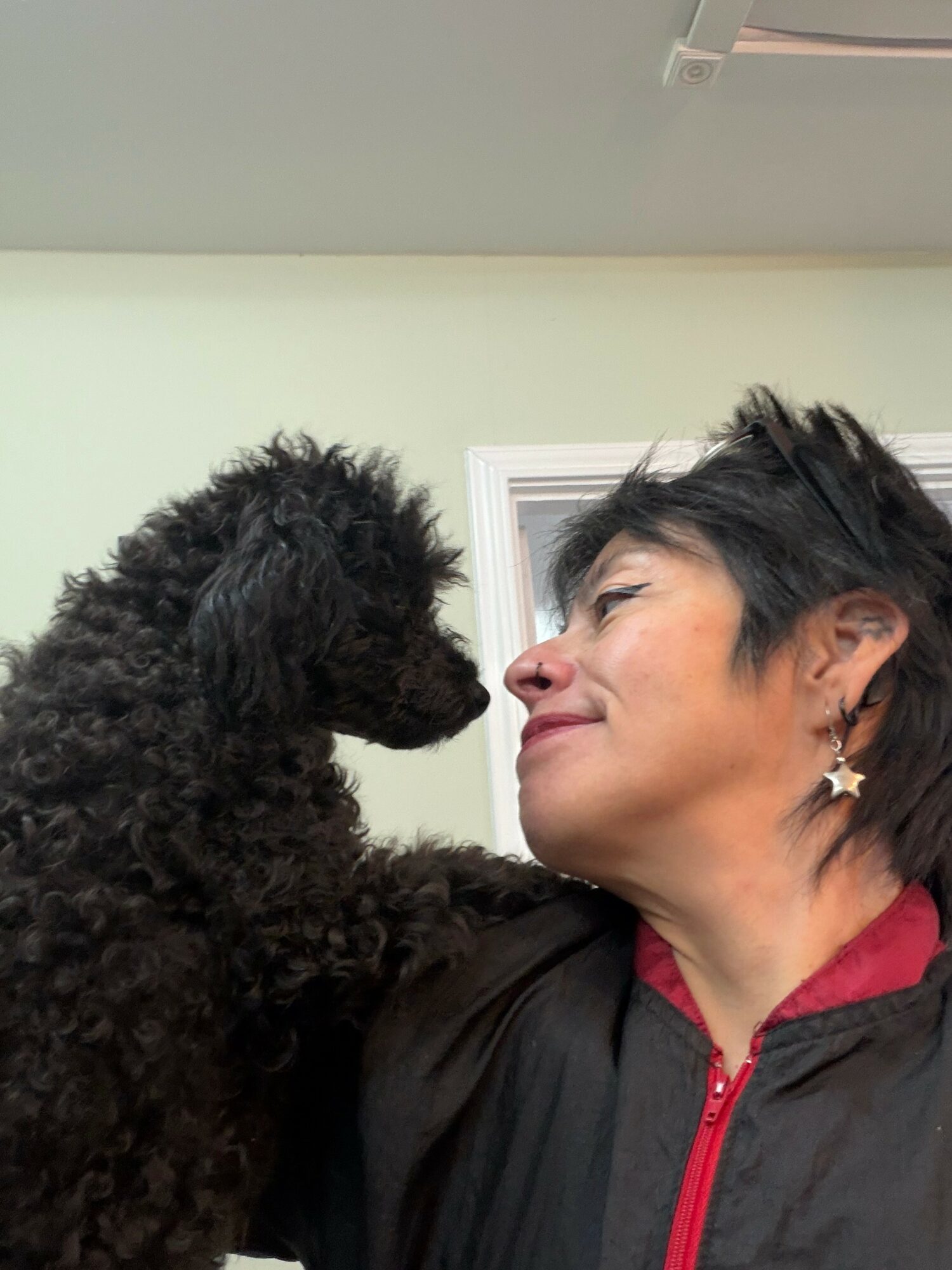 Person with short dark hair smiling at a black poodle close to their face indoors.