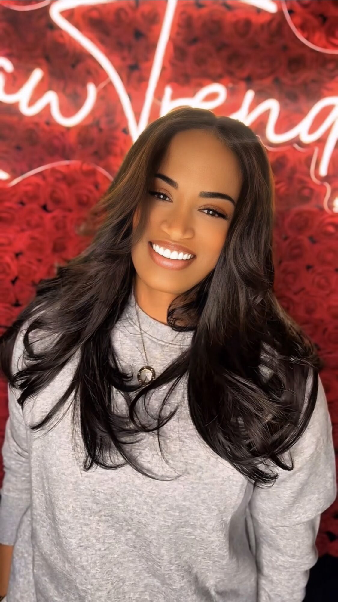 Smiling woman with long dark hair in front of a red heart background and neon sign, wearing a gray sweatshirt.