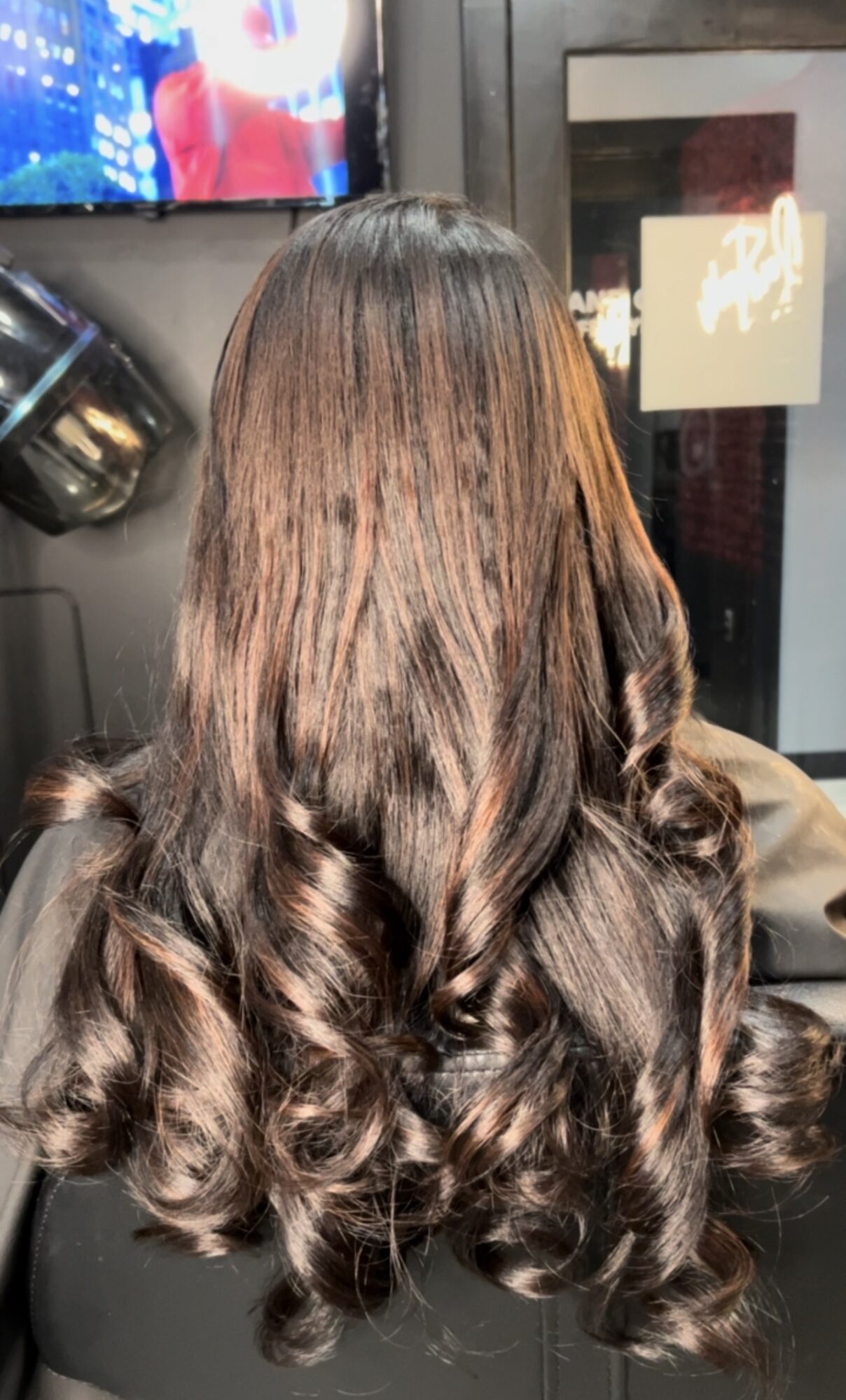Back of a person with long, wavy brown hair, indoors near a window and a television screen.